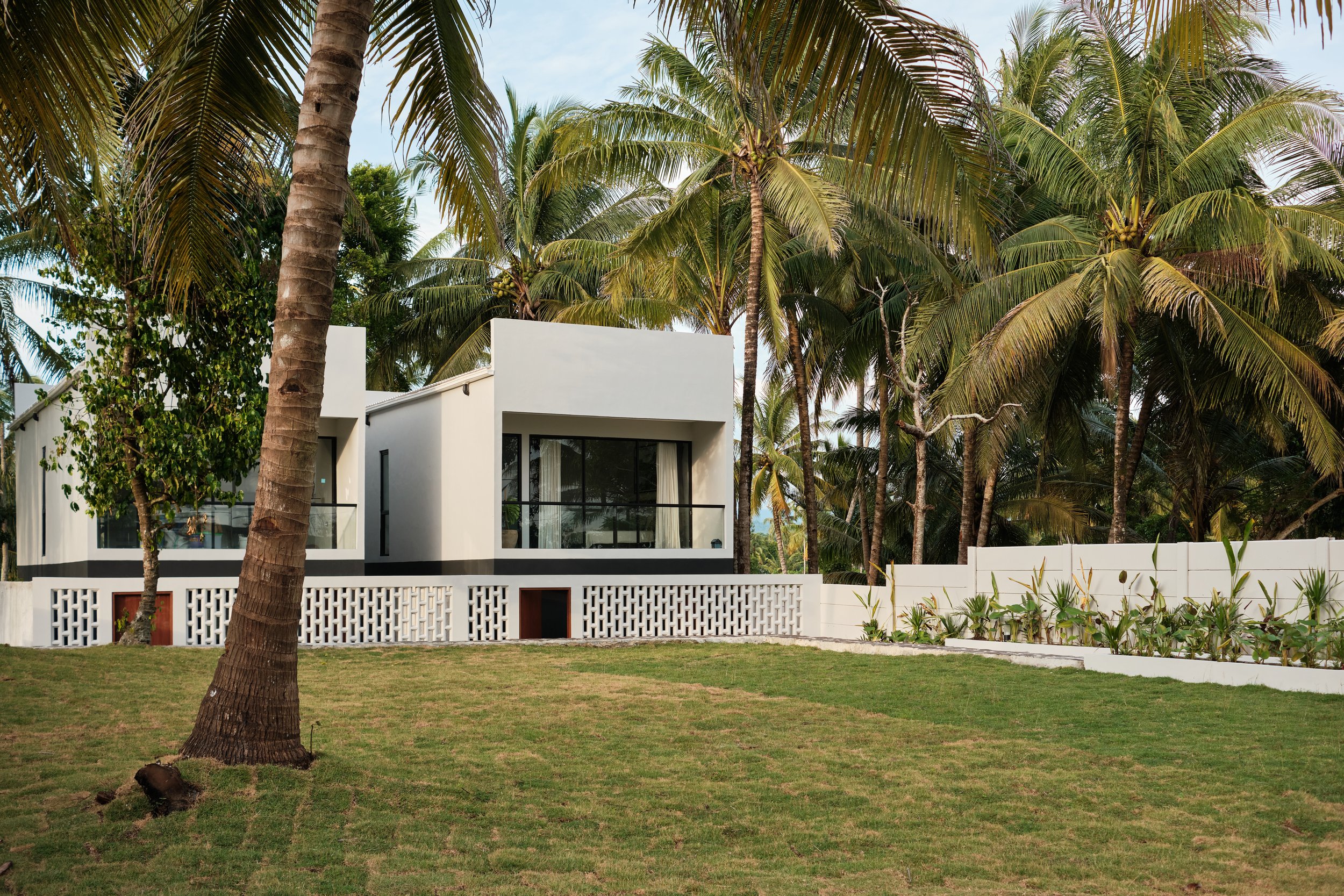 Modern white house with large glass windows, surrounded by lush green grass and tall palm trees.
