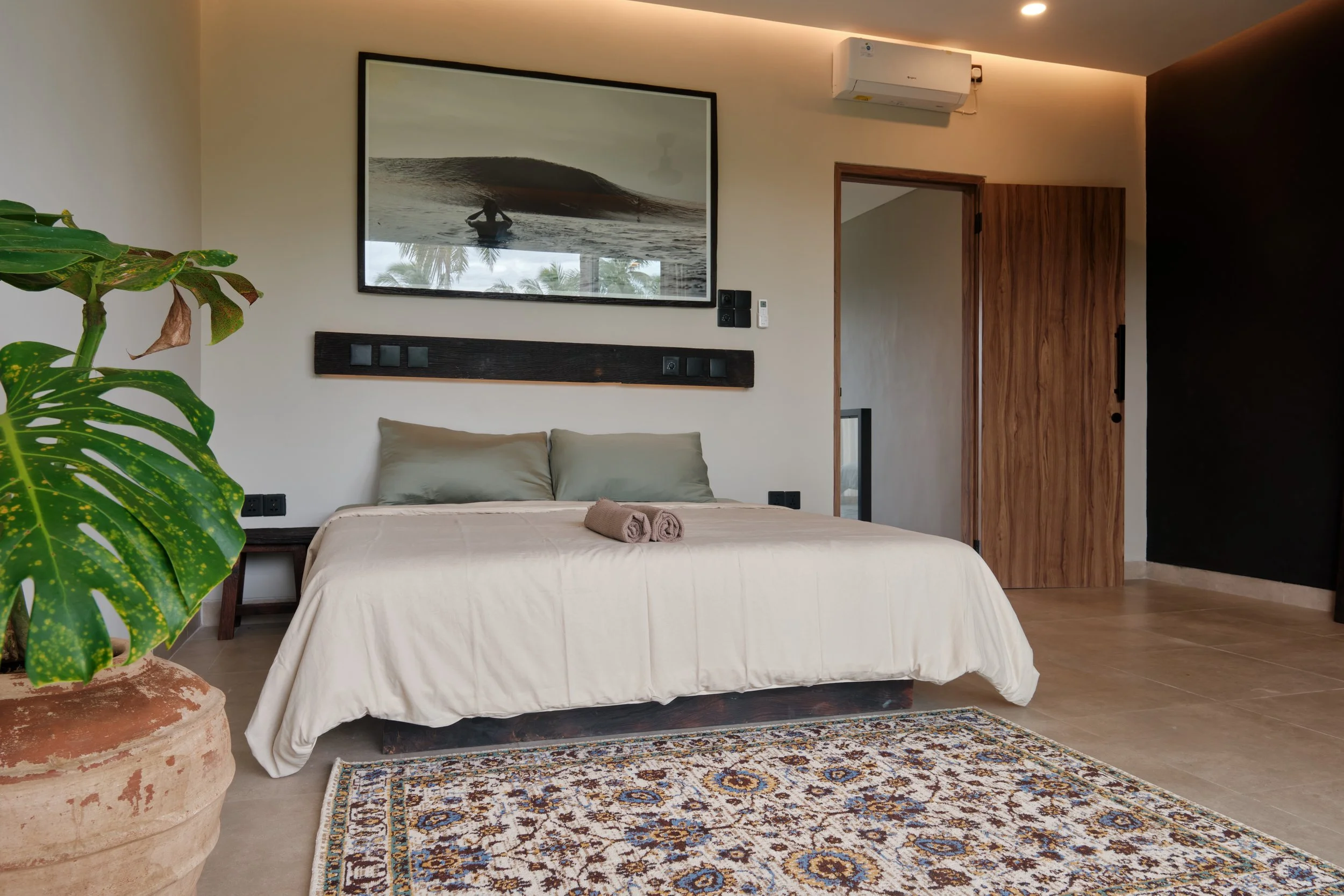 A bedroom with a large bed covered in white bedding and a pair of beige pillows. Two neatly rolled towels sit on the bed. To the left, there is a green potted plant with large, variegated leaves. Above the bed, a large framed black-and-white photograph of a wave and a surfer is hung on the wall. Next to the photograph, there is a wall-mounted air conditioning unit and a control panel. A wooden door is partly open, revealing an adjacent room or hallway. The floor features a patterned area rug, and the walls are painted in light and dark contrasting colors.