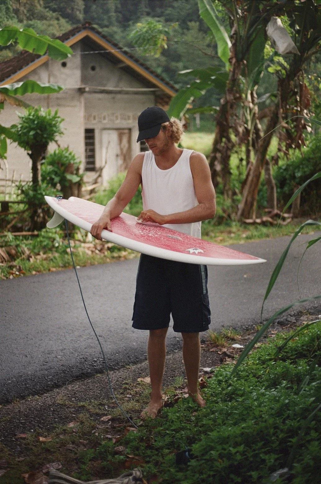 A person with curly hair, wearing a black cap, white sleeveless shirt, and black shorts, is barefoot and holding a pink and white surfboard by a tropical road surrounded by lush green plants.