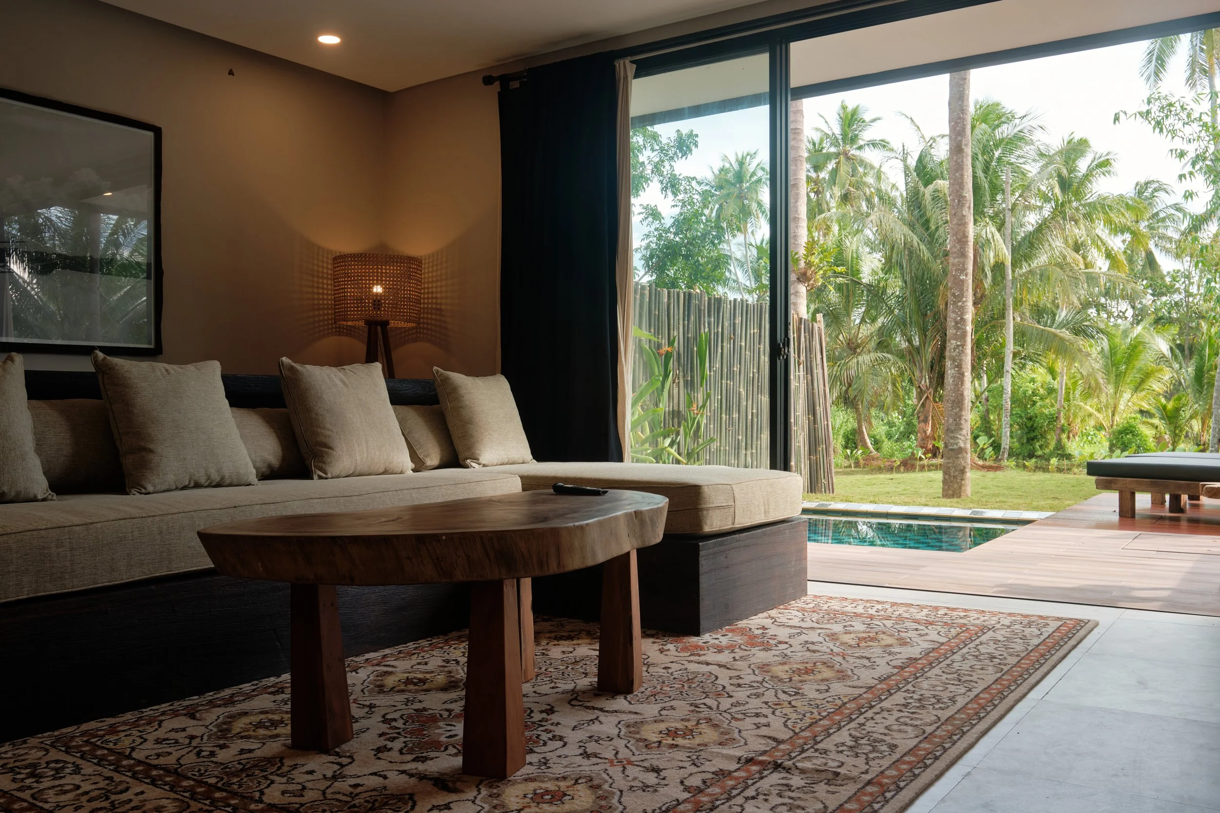 Living room with beige couch, wooden coffee table, patterned rug, floor lamp, large sliding glass door showing tropical plants and trees outside.