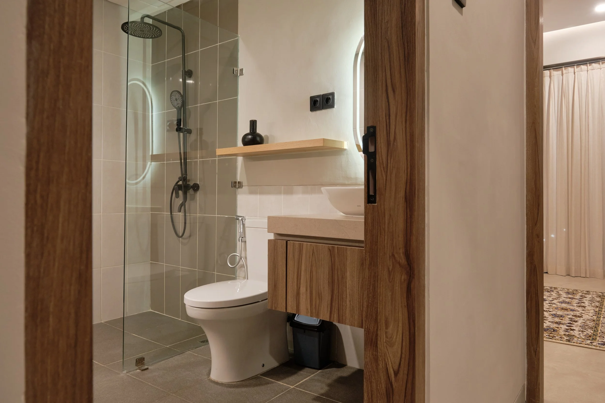 A modern bathroom with a walk-in shower, a wall-mounted toilet, a wooden vanity, and a mirror with lighting. The bathroom has neutral tones with tiles on the floor and shower wall, and a small decorative vase on the shelf.