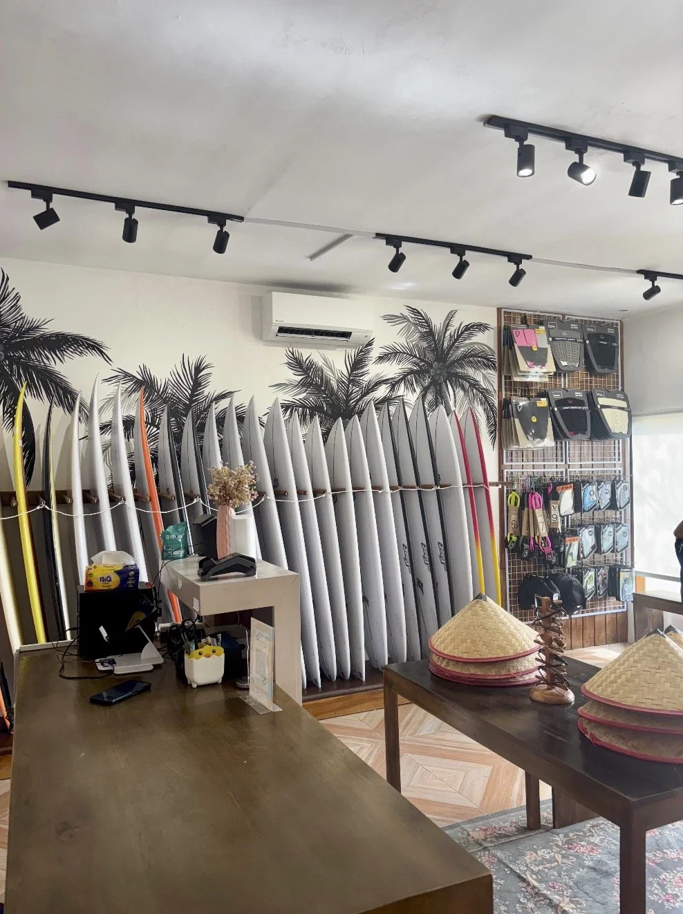 Interior of a surf shop with surfboards lined up against the wall, palm tree wall art, and racks of surf accessories and fins.