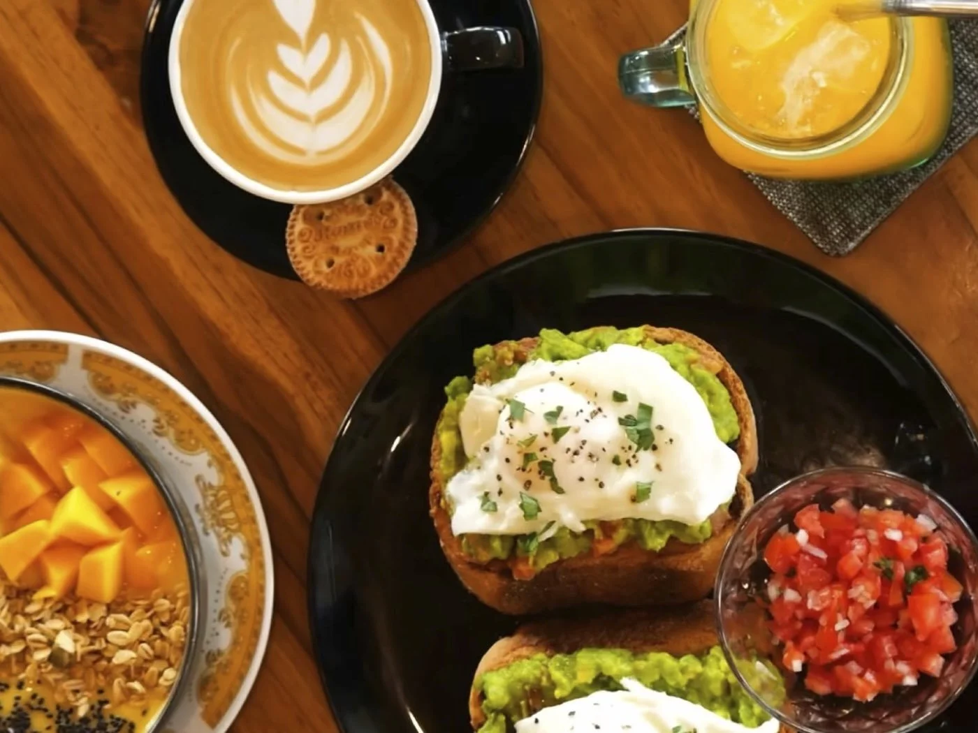 Top view of a breakfast spread with avocado toast topped with a poached egg, a bowl of fresh fruit, a cup of coffee with latte art, a glass of orange juice, and a bowl of diced tomatoes.