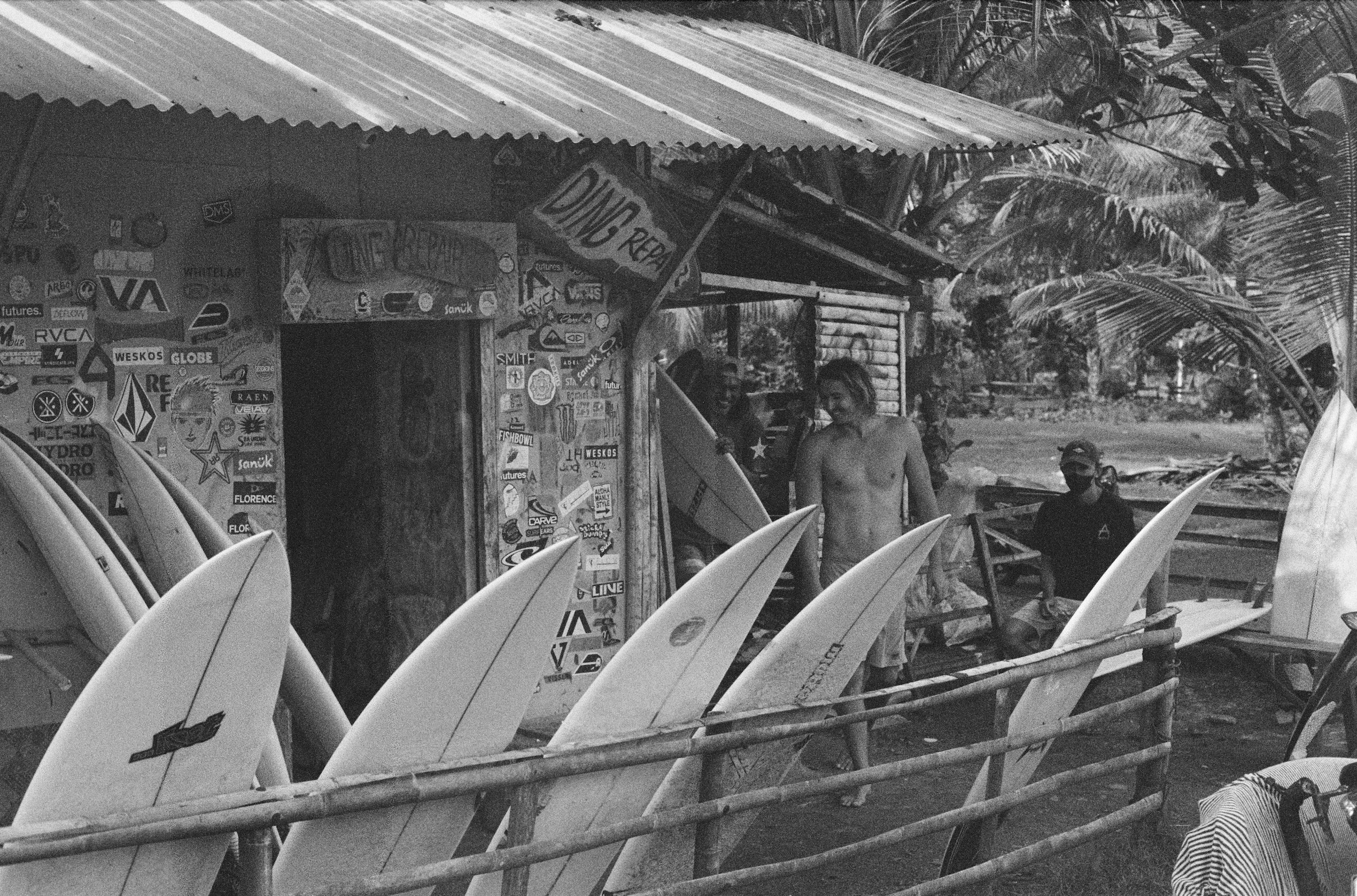 A group of surfboards lined up in front of a small building decorated with various stickers and signs, surrounded by palm trees and tropical vegetation, with two shirtless men standing nearby under a corrugated metal roof.