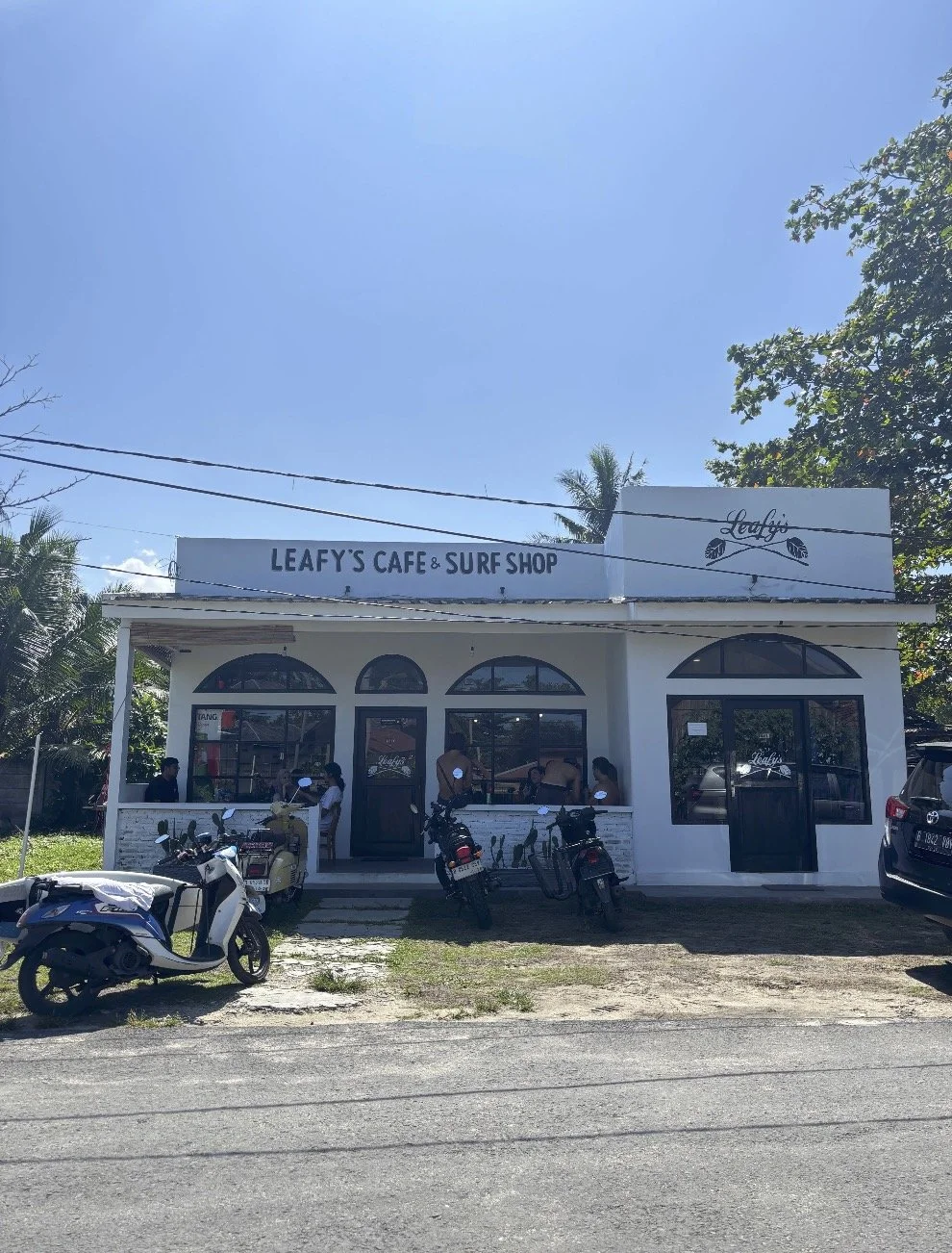 White building with signs reading 'Leafy's Cafe & Surf Shop' and 'Leafy'; large arched windows and door; motorcycles parked in front; a few people outside the cafe; sunny weather with clear blue sky.
