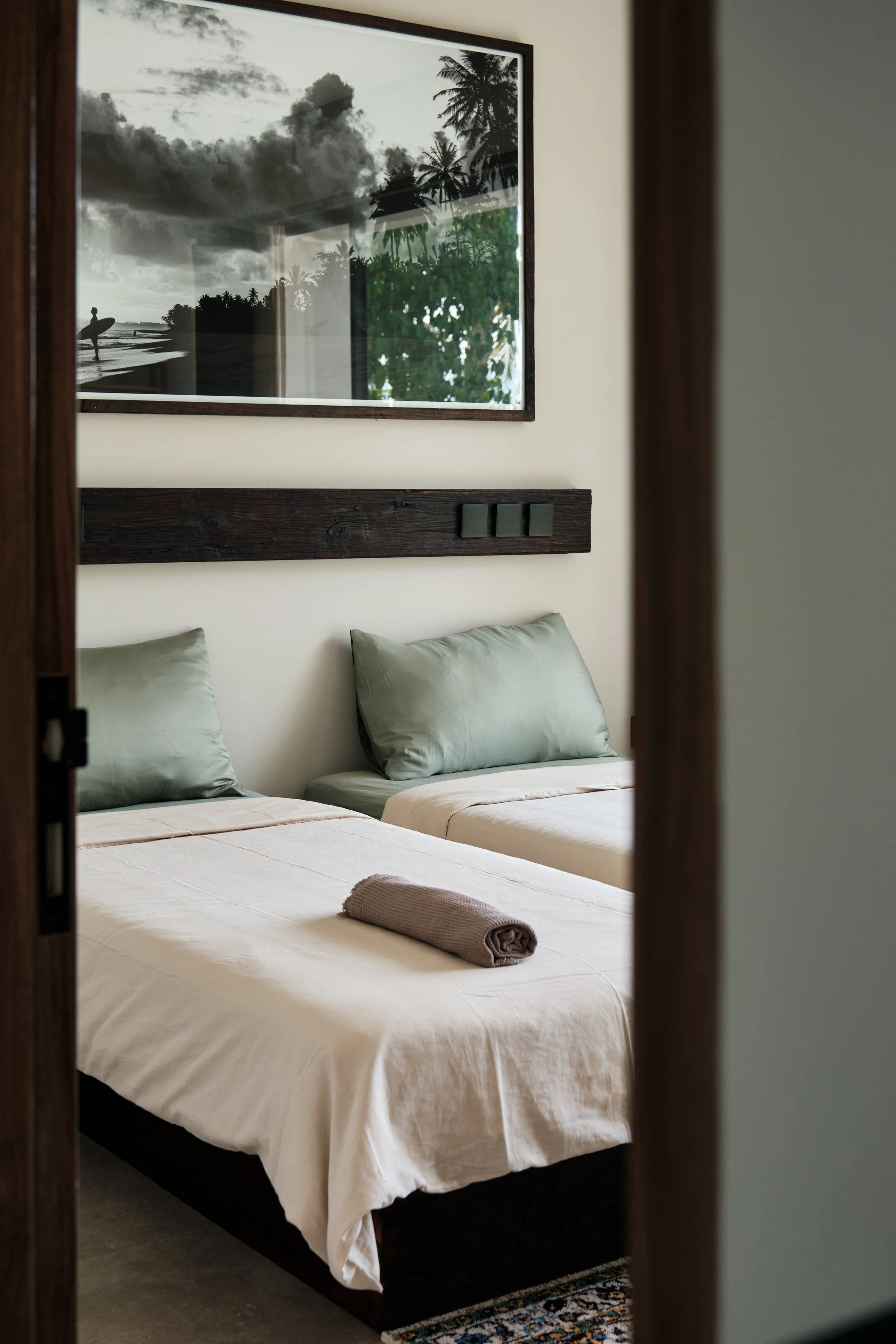 A bedroom with two beds, each with green pillows and white bedding. On the bed closer to the viewer, there is a rolled beige towel. A large picture of a tropical beach scene with palm trees, clouds, and a person holding a surfboard is hanging on the wall above the beds. The photo is reflected in a mirror on the left side of the image.