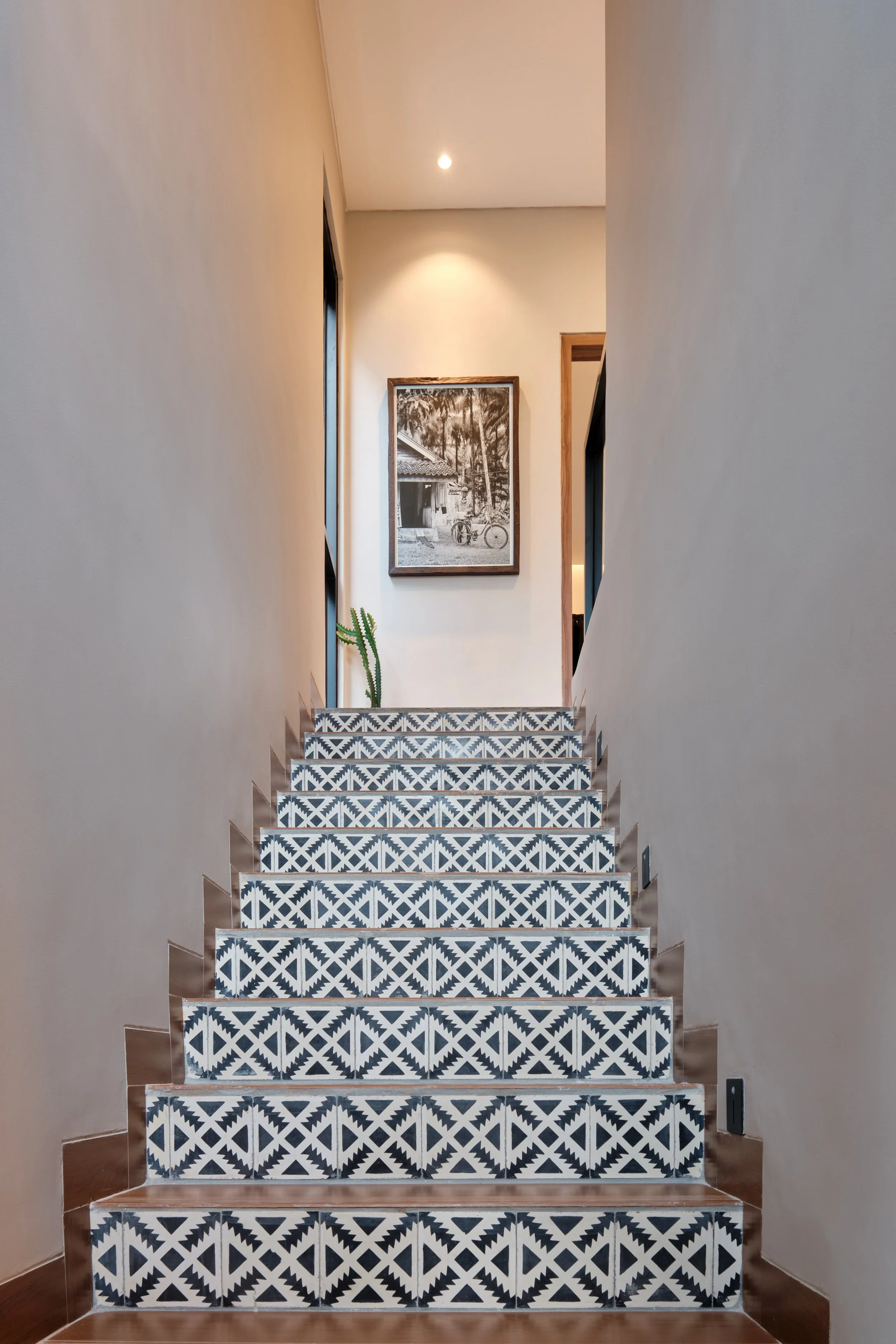 Indoor staircase with black and white patterned tiles, beige walls, framed black and white photograph of trees and bicycles at the top, and a potted plant on the right.