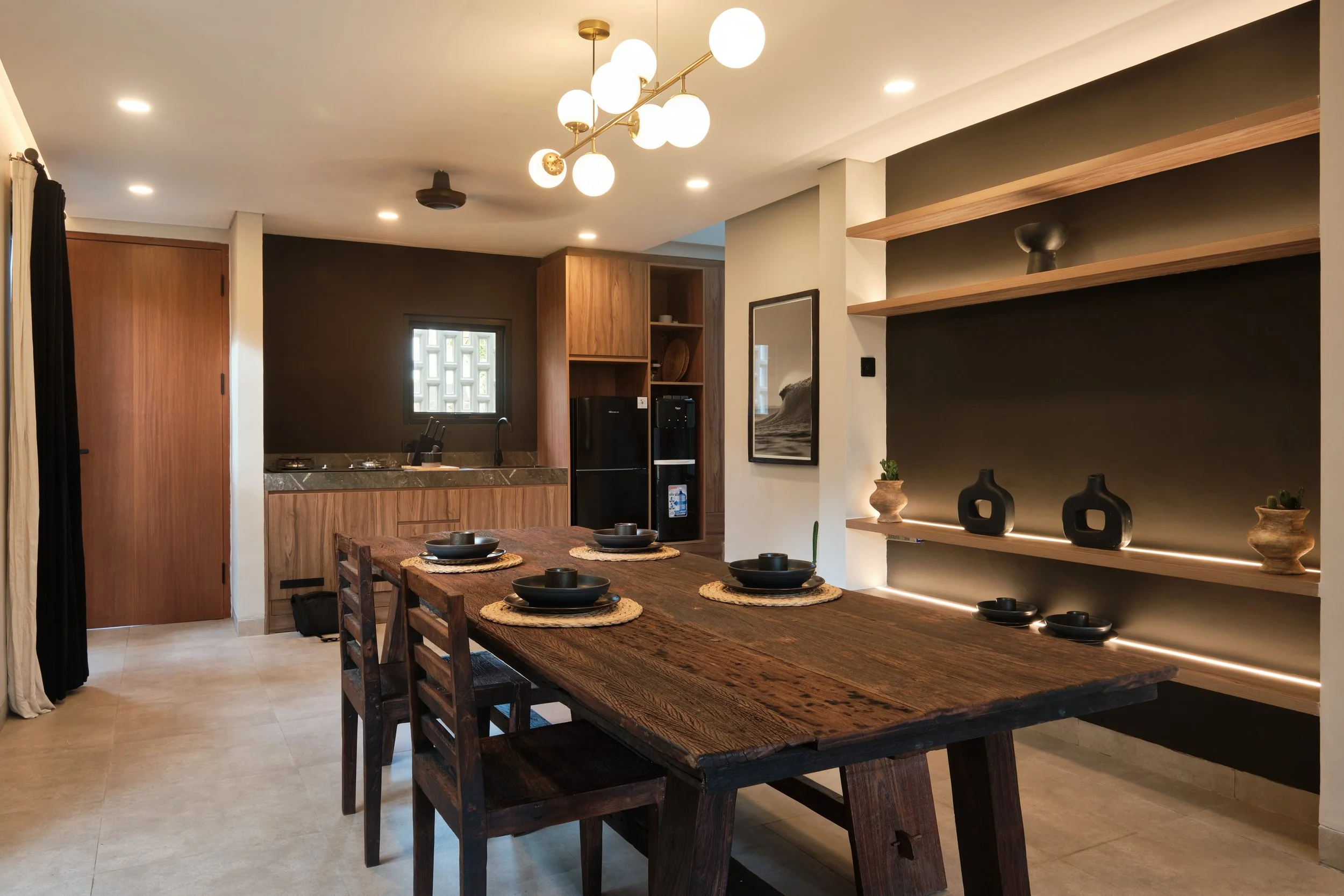 Interior view of a modern dining and kitchen area with wooden furniture, black decorative vases, and built-in shelving with lighting.