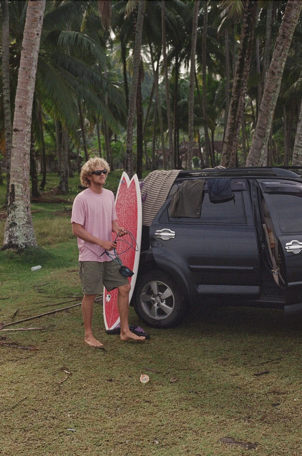 A man with curly blond hair wearing sunglasses, a pink t-shirt, and khaki shorts stands on a grassy area holding a surfboard and a leash. He is barefoot and next to a black SUV with its rear door open, with clothes hanging from the vehicle. The background features tropical trees and greenery.