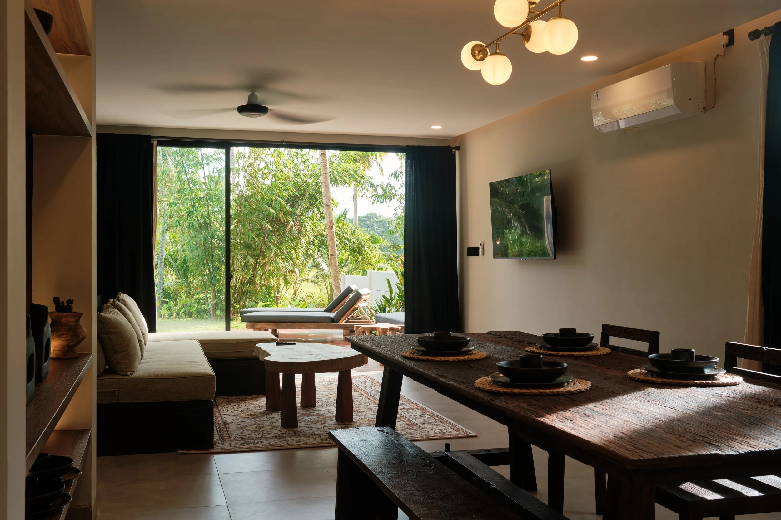 A living room with large sliding glass doors opening to a lush green outdoor area, featuring lounge chairs. The room has a ceiling fan, modern lighting, a wall-mounted TV, an air conditioner, and a dining table set with black bowls on woven placemats.