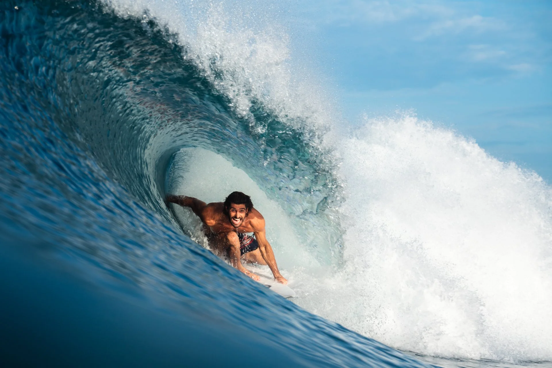 A man with dark hair and a beard surfing on a blue ocean wave with a clear blue sky in the background.