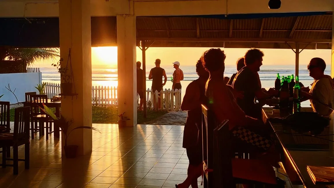 People enjoying drinks at a beachside restaurant during sunset, with the ocean and sky visible in the background.