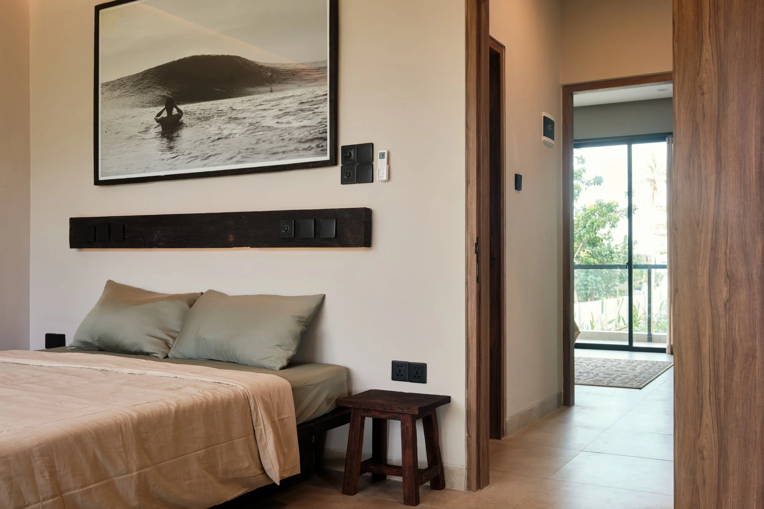 A bedroom with a bed, a black headboard, beige pillows, and a beige bedspread. There is a wooden stool beside the bed and framed black-and-white photo of a surfer riding a wave on the wall behind the bed. A window with a sliding door shows greenery outside.