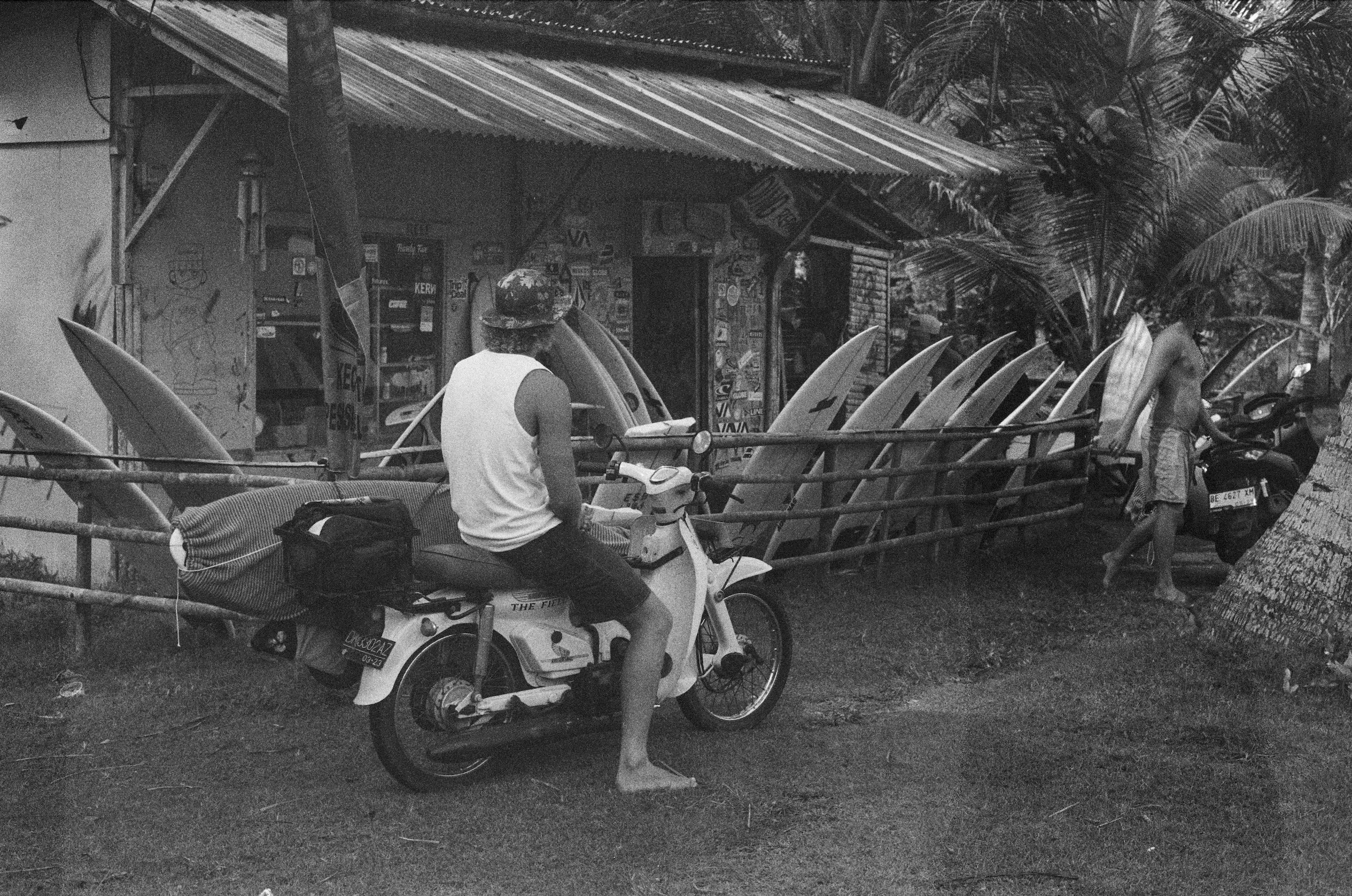 A black and white photo of a small shop with surfboards lined up outside. Two men are near a scooter and a woman in a skirt walks past in a tropical setting with palm trees.