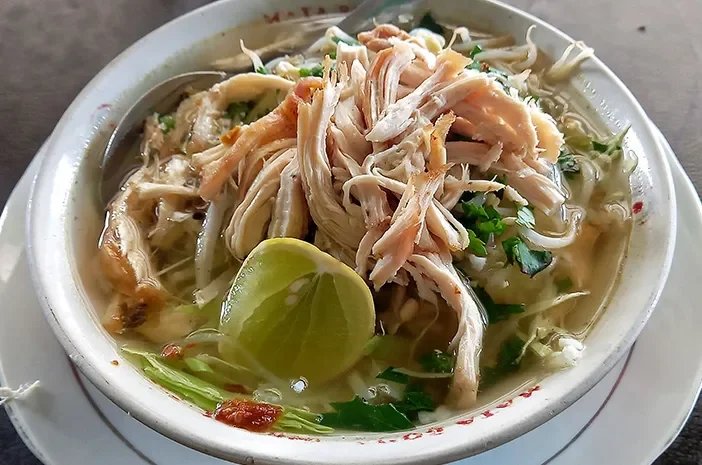 A bowl of chicken noodle soup with shredded chicken, lime wedge, cilantro, and noodles in broth.