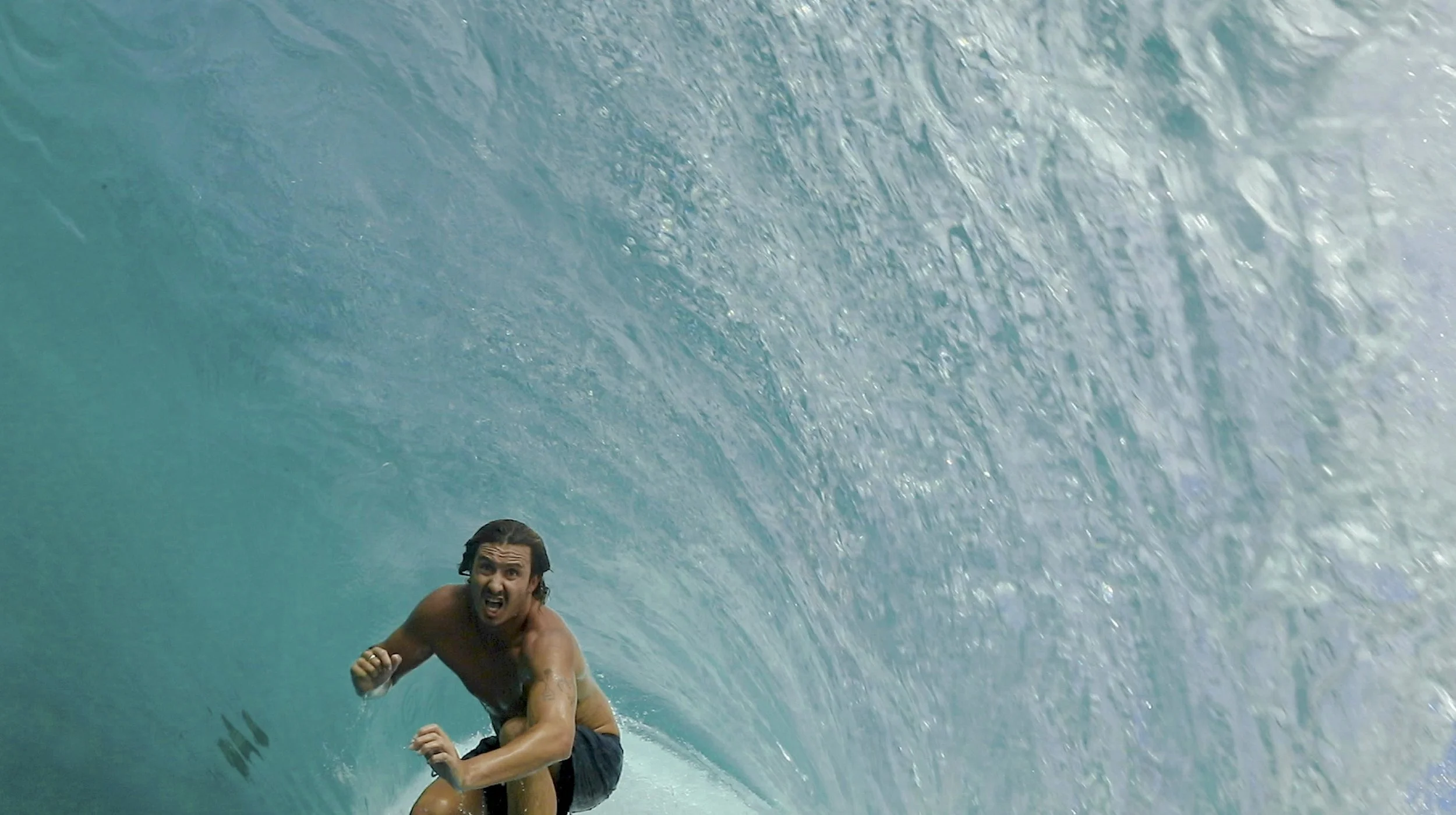 A man with long dark hair and a beard surfing inside a large blue ocean wave, with an expression of excitement and determination.