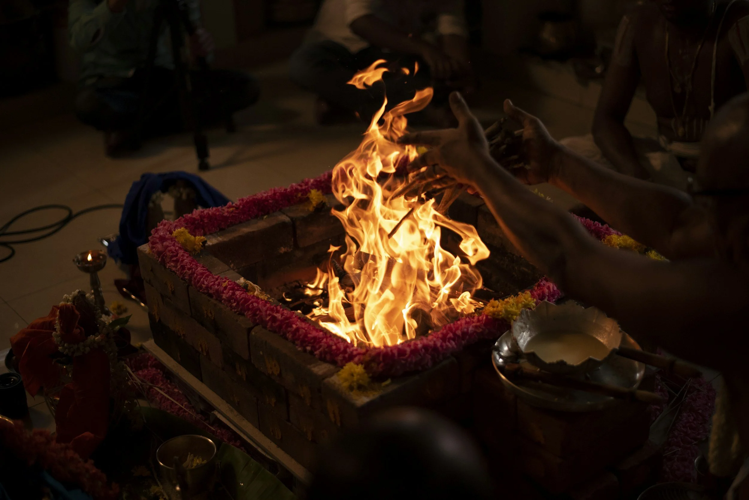 People performing a traditional fire ritual with offerings, surrounded by flowers, in a religious or spiritual setting.