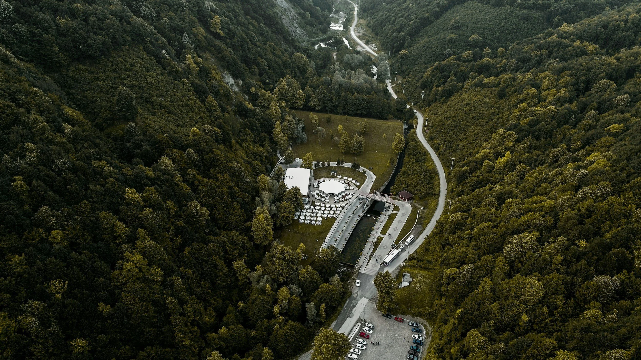 Aerial view of a scenic area with winding roads, a small parking lot, and a structure with outdoor seating surrounded by dense green forest and mountain hills.