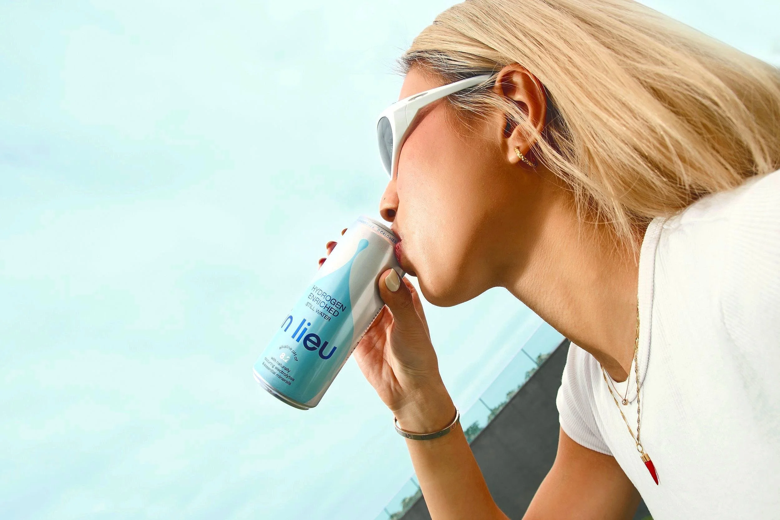 A woman with blonde hair, wearing sunglasses, earrings, and layered necklaces, drinking a can of in lieu hydrogen-enriched still water outdoors.