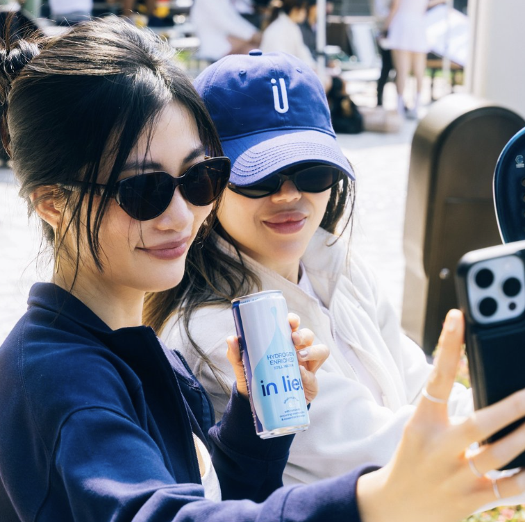 Two women wearing sunglasses and hats taking a selfie outdoors, with one holding a can of in lieu hydrogen-enriched still water in a blue and white can.