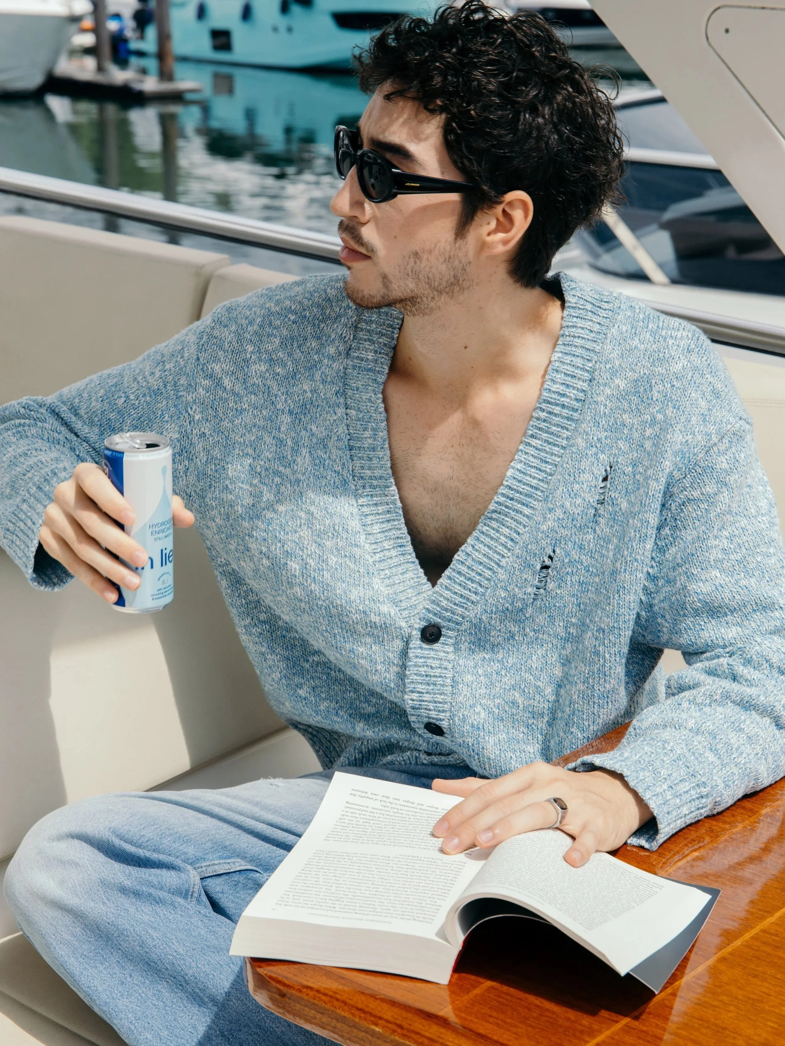 A man sitting on a boat, wearing sunglasses and a blue cardigan, holding a can of in lieu sparkling water, with an open book on his lap, near a marina with boats in the background.