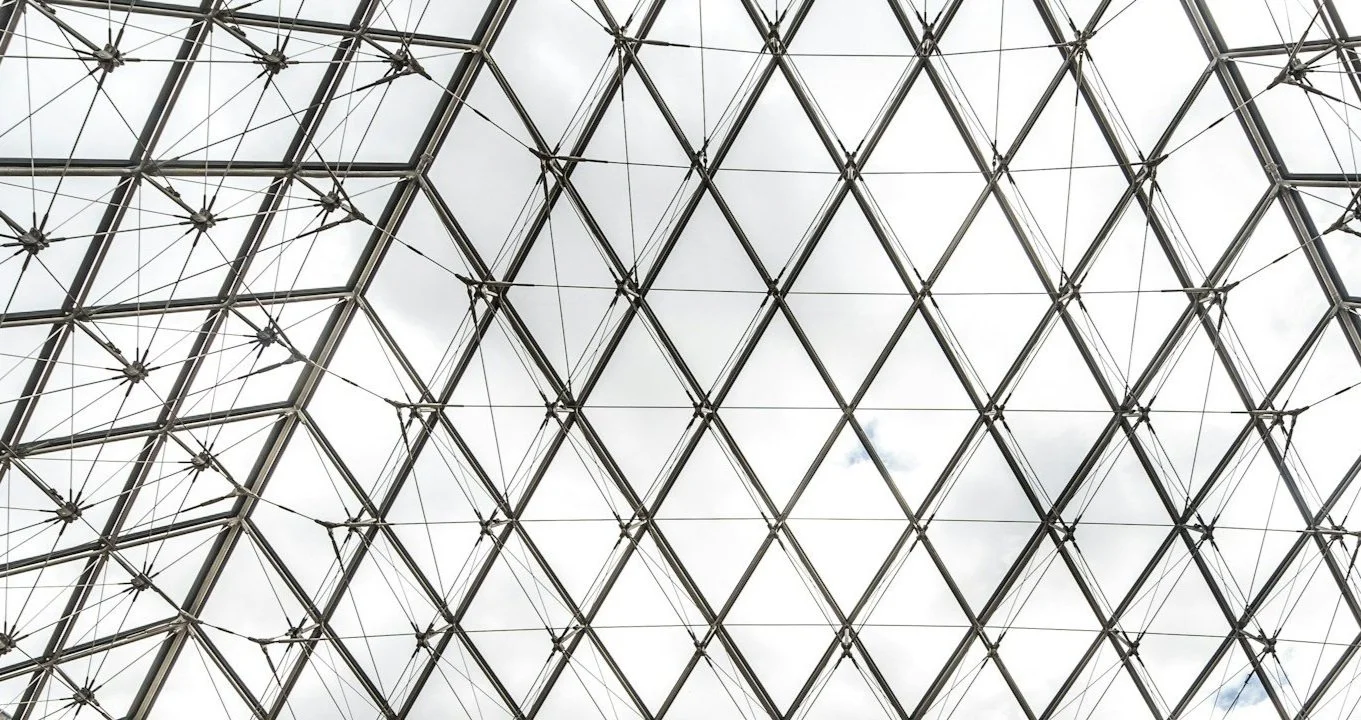 Looking up at a large glass and metal geometric dome ceiling structure with a cloudy sky visible through the glass panels.