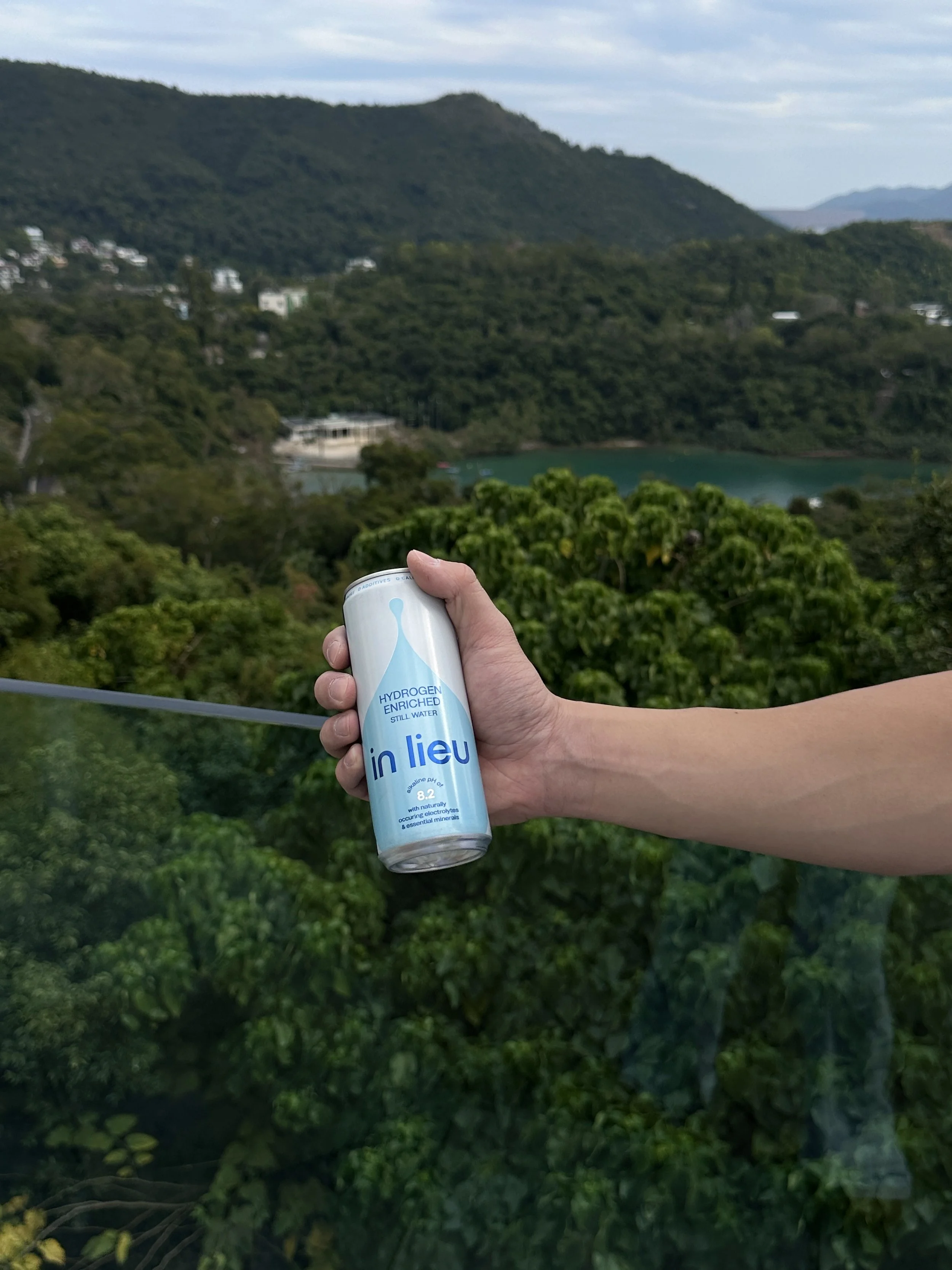 A person holding a can of in lieu hydrogen-enriched still water in front of a lush green landscape with hills, trees, and a lake.