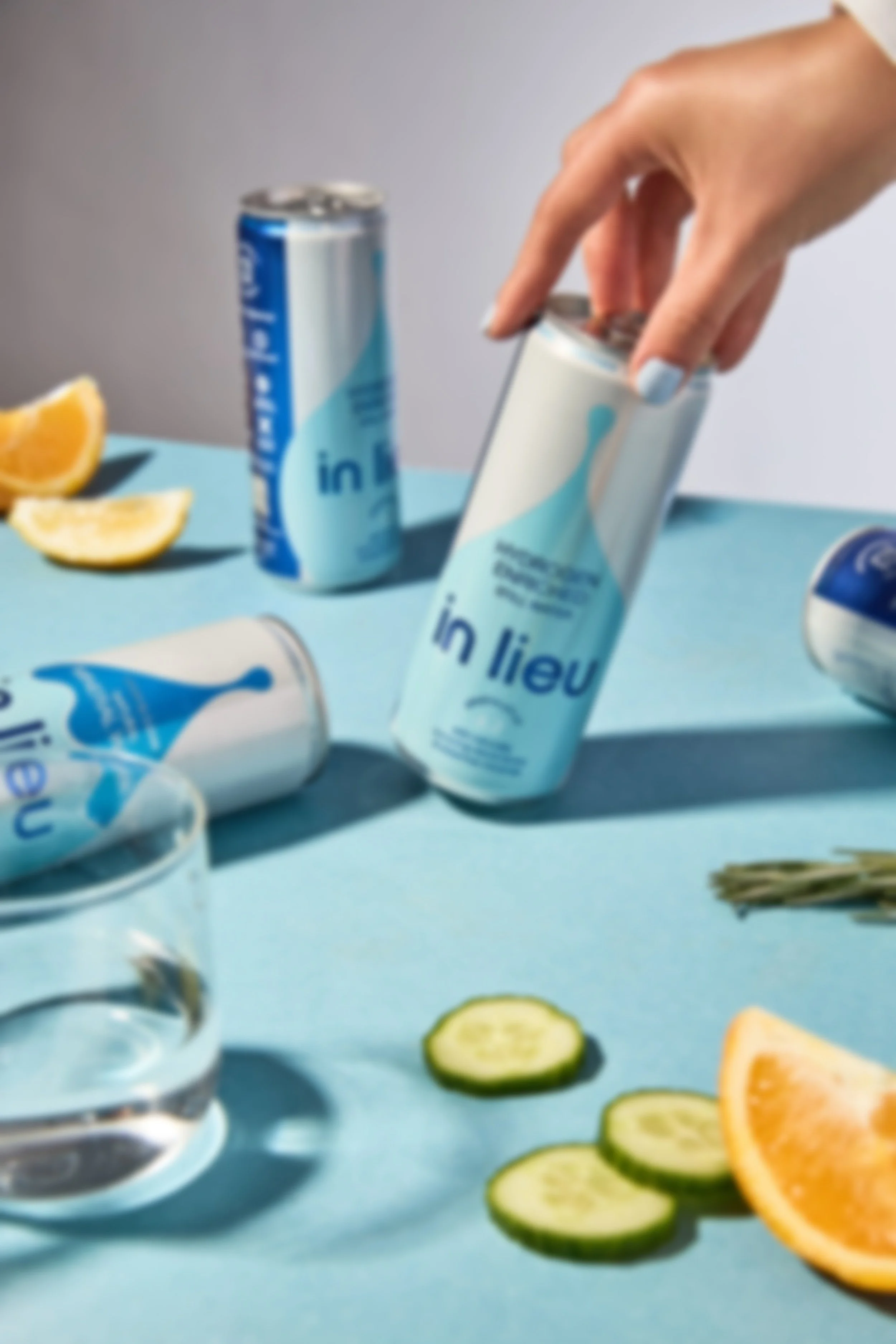 A hand reaching to pick up a can of in lieu hydrogen-enriched still water, with other cans, sliced cucumber, orange, and a glass of in lieu water on a blue surface.
