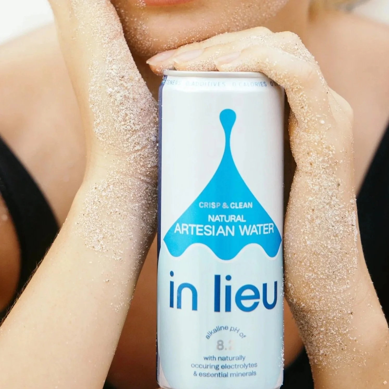 Person with sand on hands and arms holding a can of in lieu natural artisanal water near face.