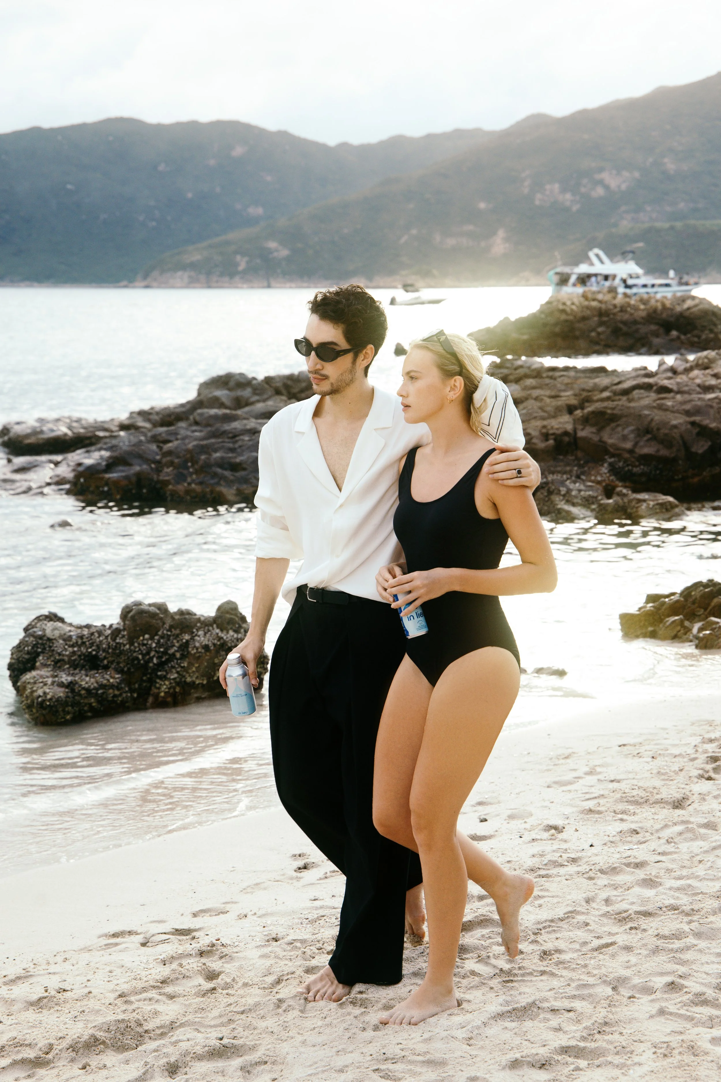 A young man and woman walk along a sandy beach with rocks and the ocean in the background. The man wears sunglasses, a white shirt, and black pants, while the woman wears a black swimsuit. They both hold bottles of water.