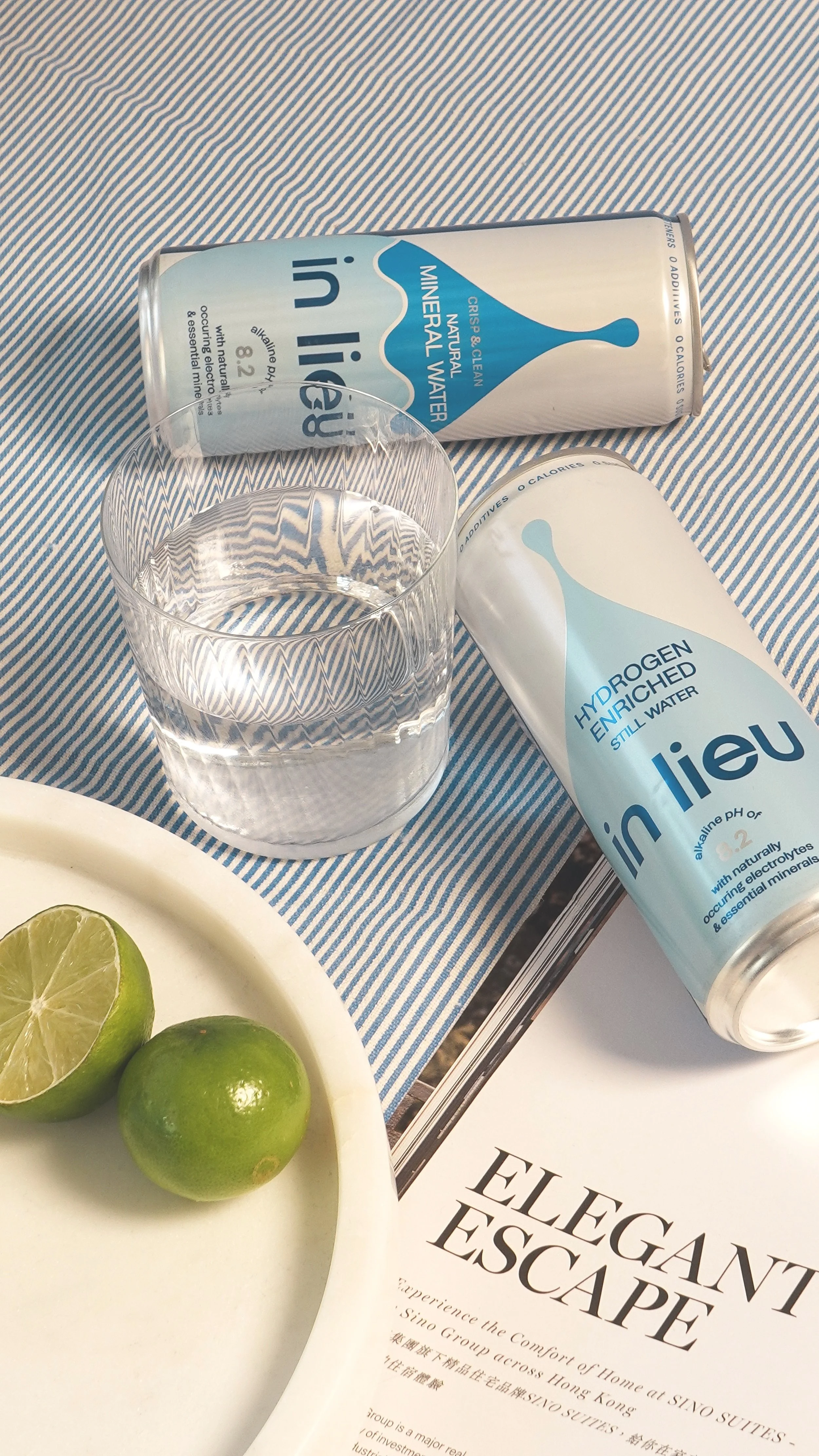 A glass of clear mineral water on a striped tablecloth, two cans of in lieu hydrogen-enriched still water, and a white plate with a halved lime and a whole lime.