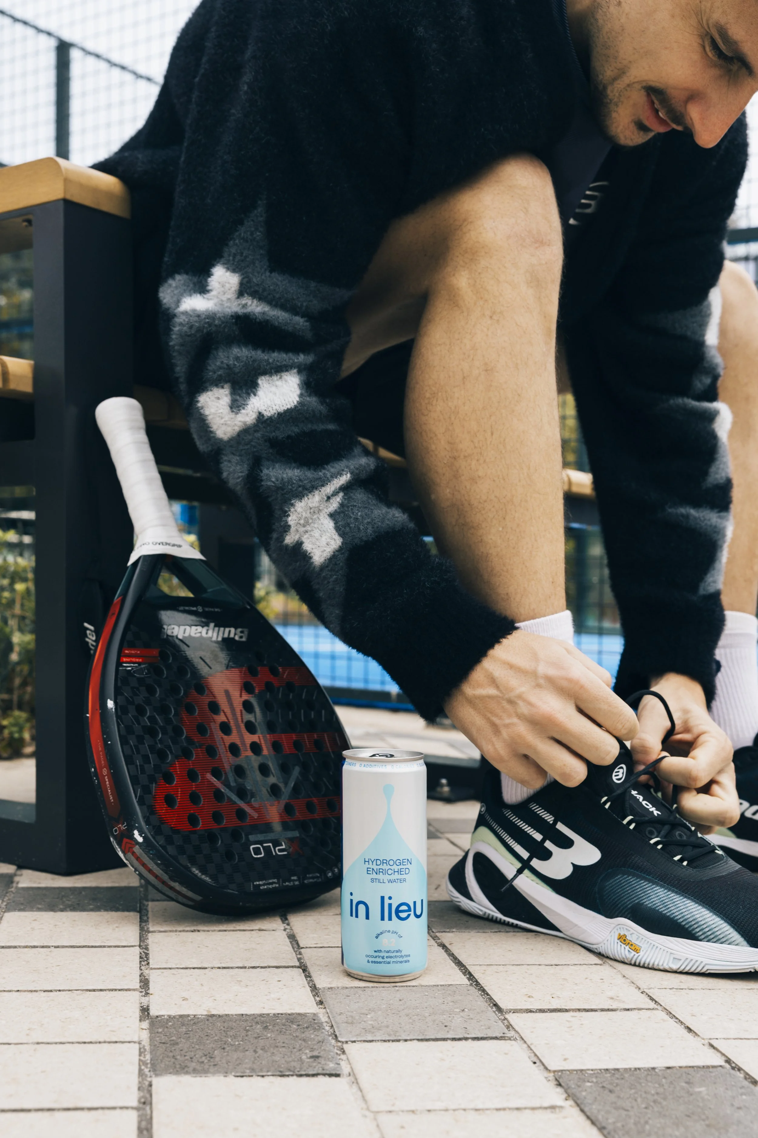 A man tying his tennis shoe next to a paddle, a can of in lieu hydrogen-enriched still water, and a bench on an outdoor tennis court.