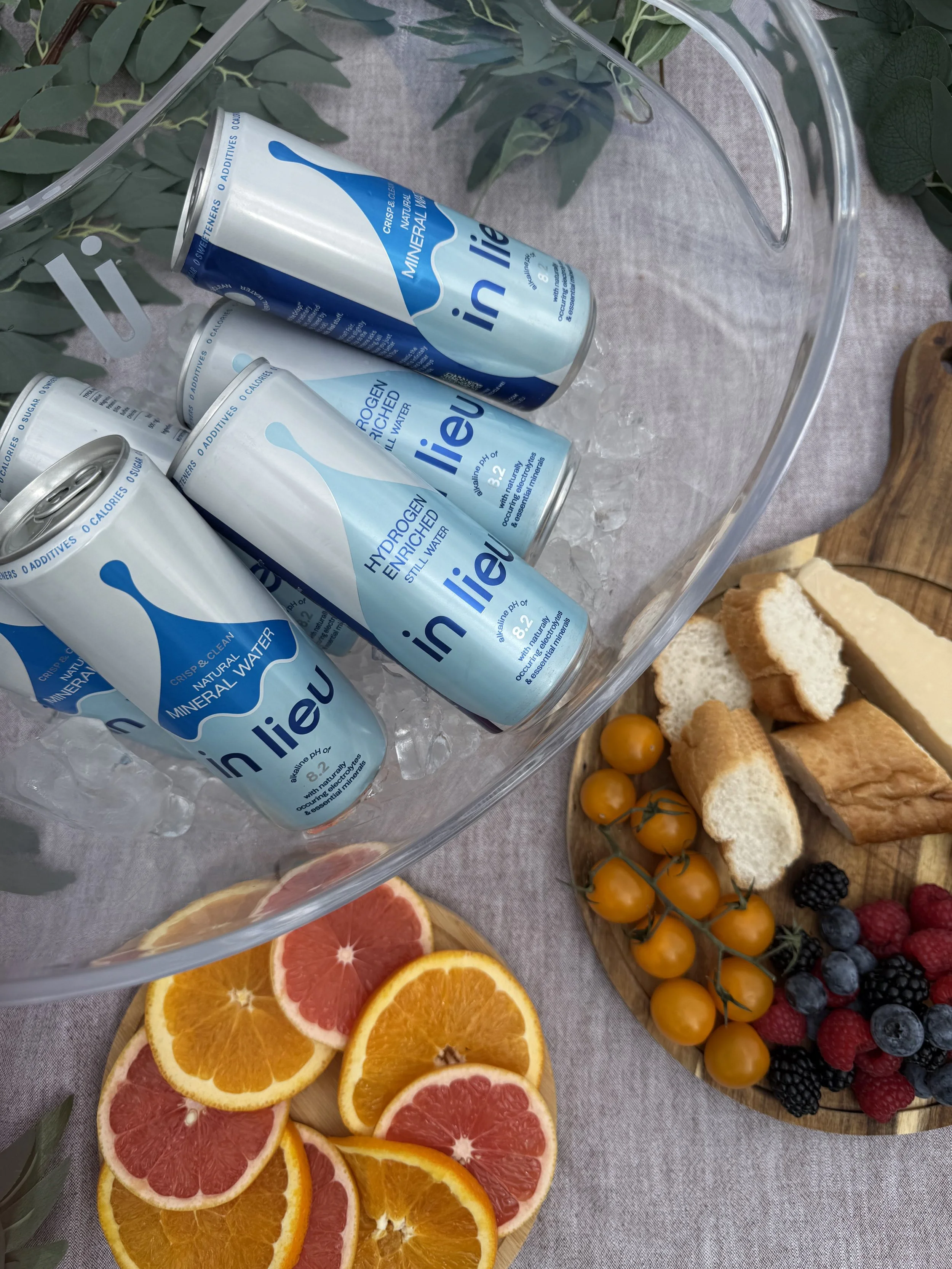 Several cans of Inlieu hydrogen-enriched mineral water in a glass bowl with ice, surrounded by citrus slices, a wooden board with cherry tomatoes, bread slices, and mixed berries, set on a fabric surface with green leaves nearby.