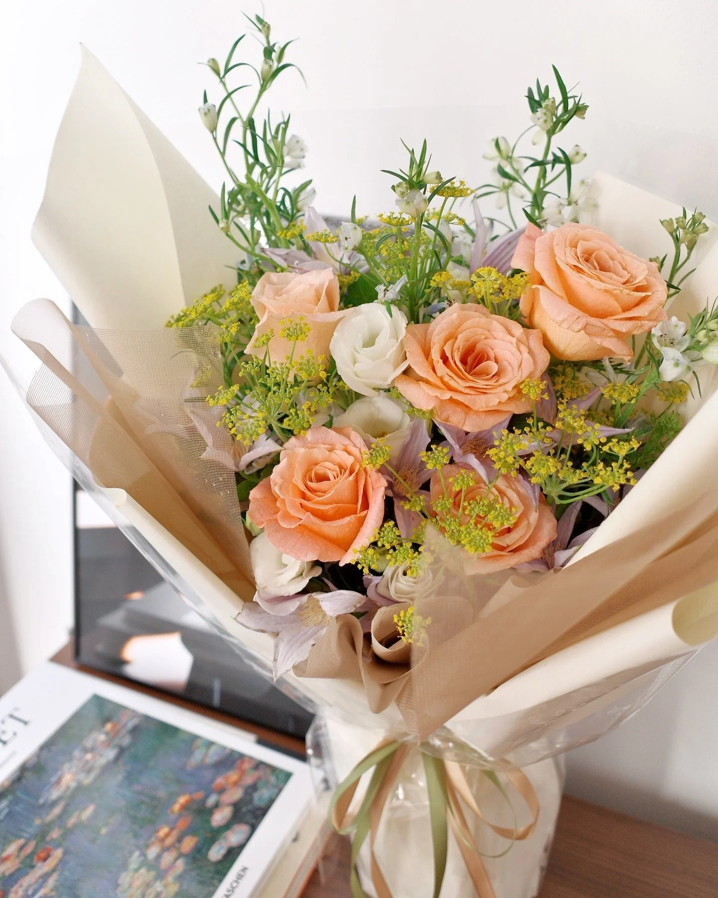 A bouquet of peach and white roses, with greenery, wrapped in beige and white paper, on a wooden surface.