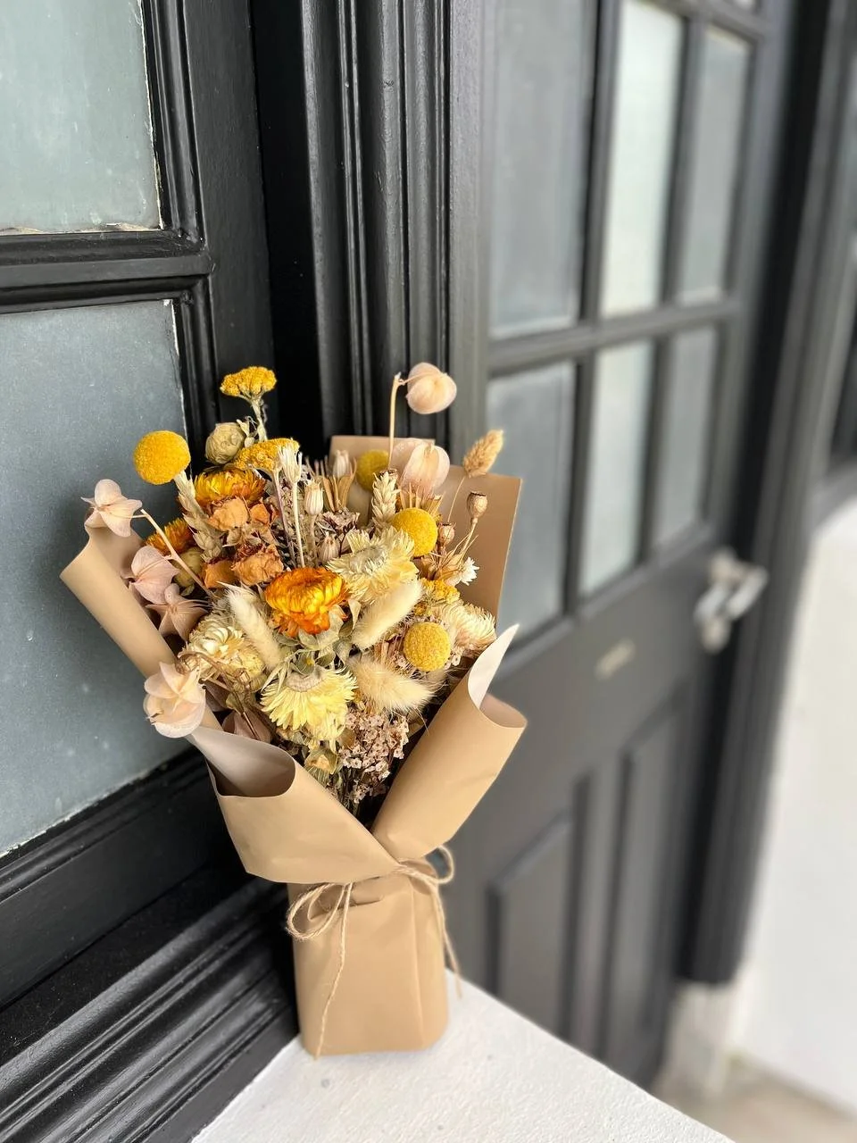 A bouquet of dried flowers, including yellow, white, and beige blooms wrapped in brown paper with a tied string, leaning against a black window frame.