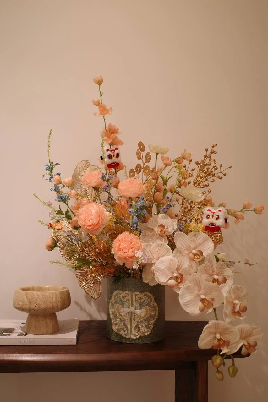 A floral arrangement in a ornate vase containing pink roses, white orchids, and various decorative elements, with two small plush lion toys among the flowers, placed on a wooden table.