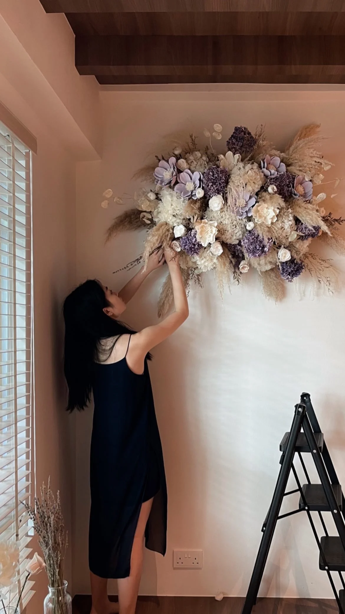 A woman in a black dress is arranging a large, elaborate floral arrangement on a wall inside a room with beige walls, a window with blinds, and a step ladder nearby.