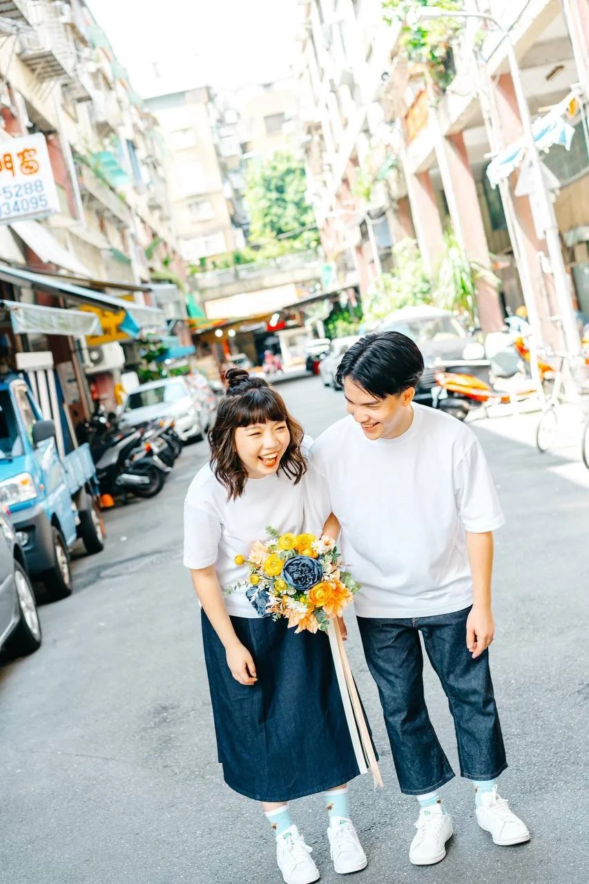 A young couple walking hand in hand on a city street, smiling and laughing. The woman is holding a colorful bouquet of flowers. The street is lined with parked cars and tall buildings.