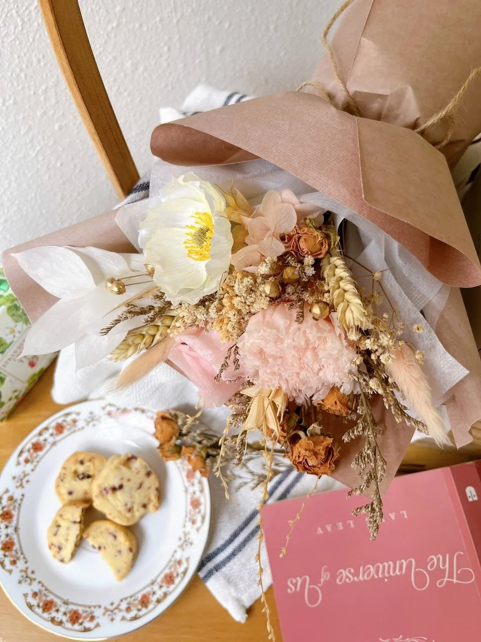 A bouquet of dried and fresh flowers in soft pastel colors, alongside a plate of berry scones on a decorative floral plate and a pink box with white text, placed on a wooden table with a striped cloth.