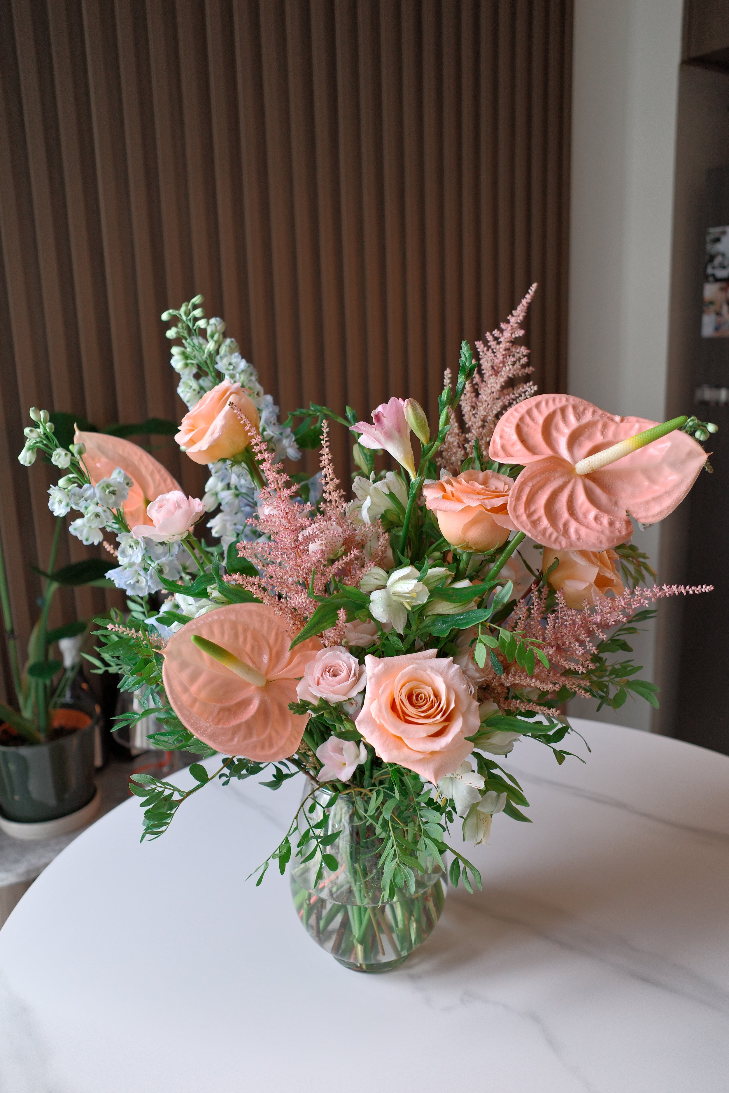 A vase with pink and white flowers, including roses, calla lilies, and anthuriums, arranged on a white table with green leaves.