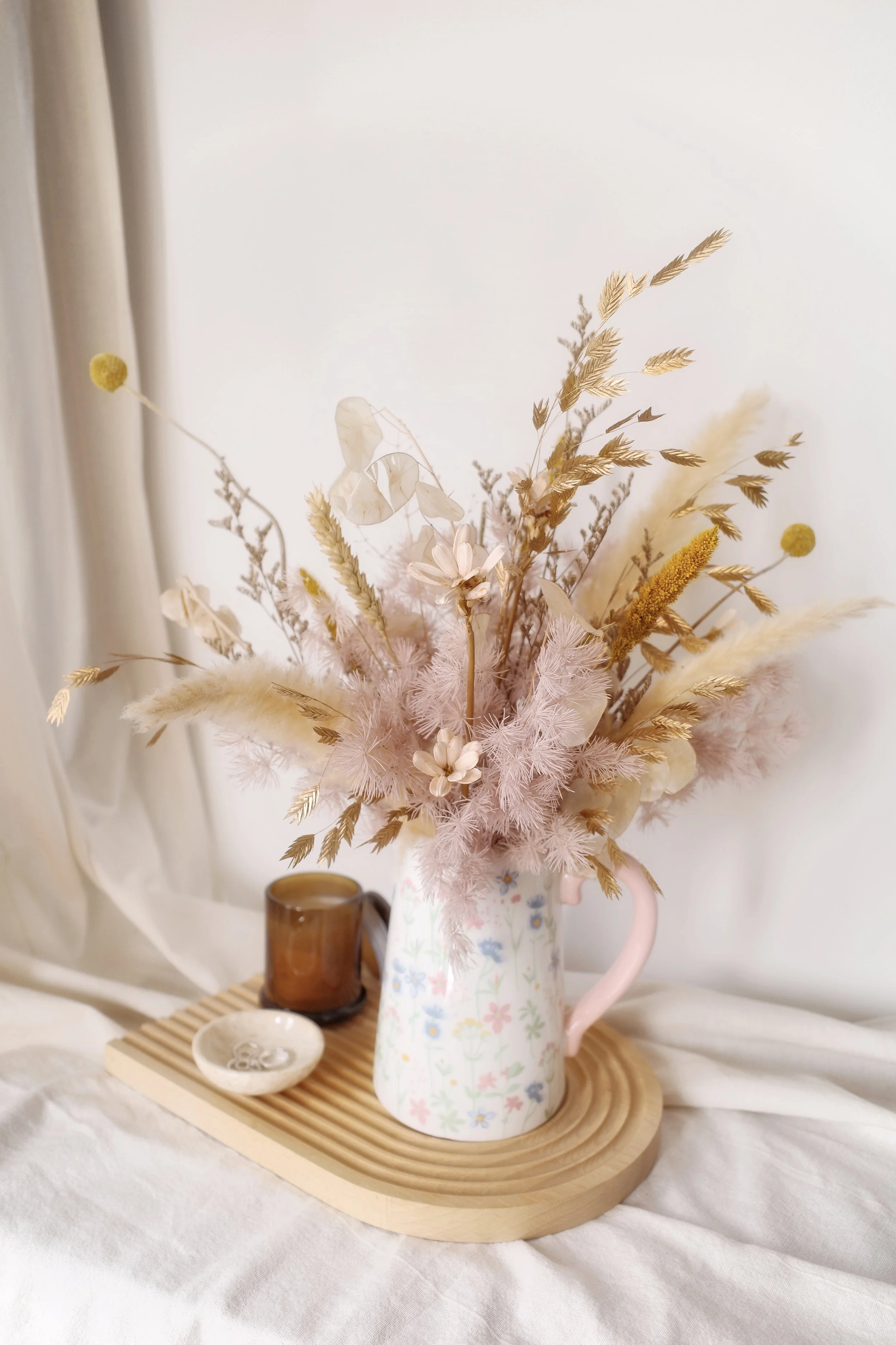 Decorative dried flower arrangement in a pastel floral pitcher on a wooden tray with a small brown mug and a white bowl, set on a bed with white sheets and near white curtains.