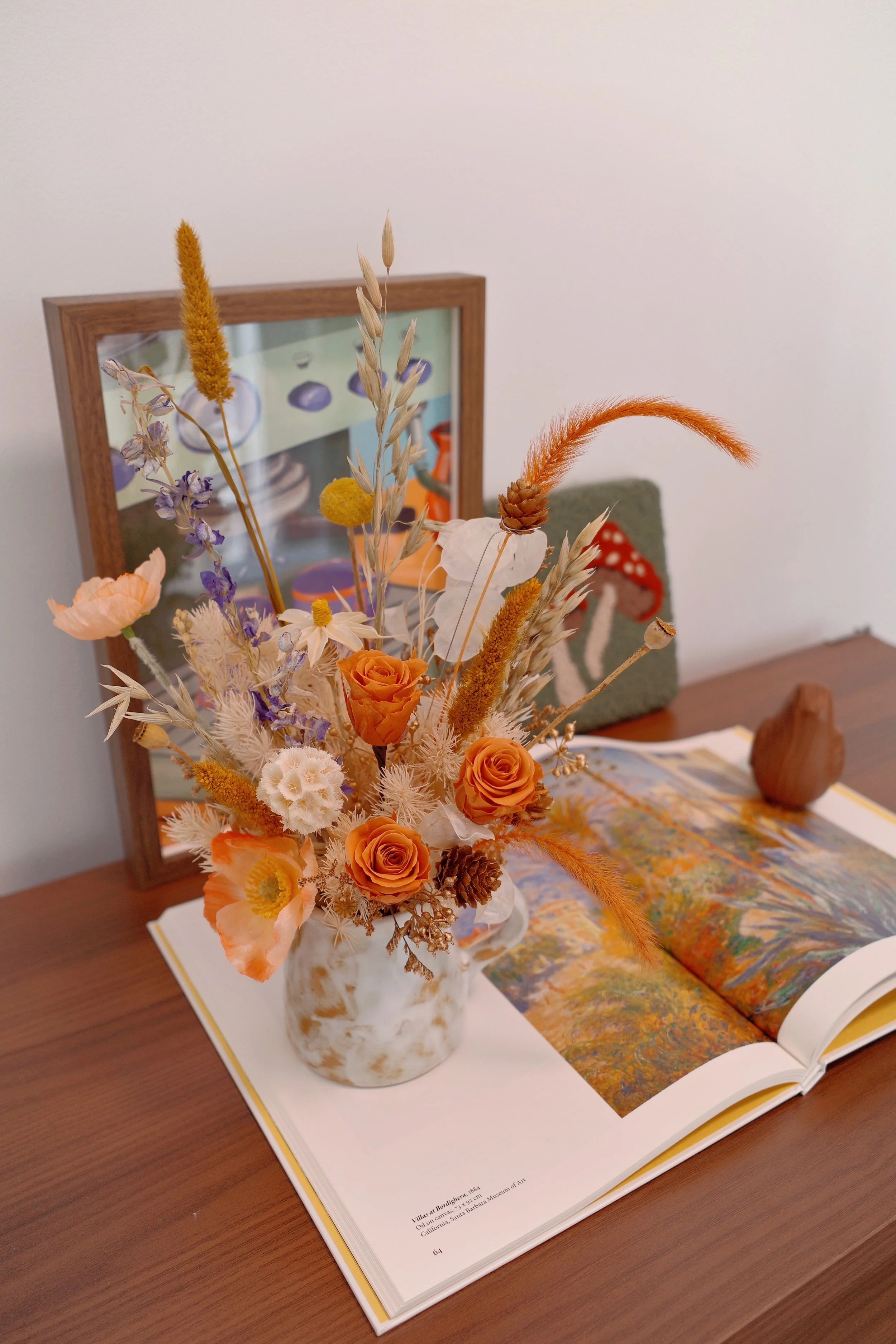 A floral arrangement with orange roses, purple and white dried flowers, and grasses in a marbled white and orange vase, placed on an open art book on a wooden table with a framed picture and a small decorative object in the background.