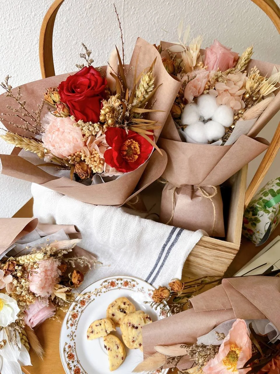 A wooden box holding bouquets of dried and fresh flowers, including roses, and a plate with four pieces of berry scone.
