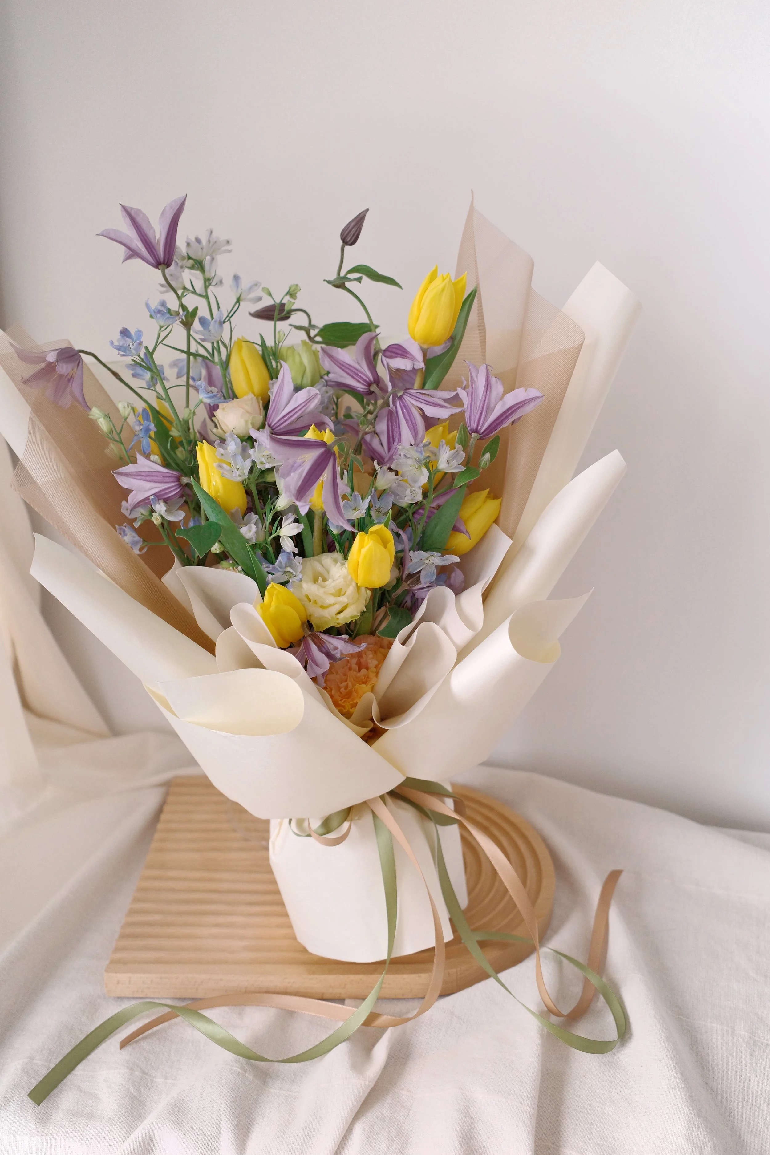 Elegant bouquet of yellow tulips and purple striped lilies wrapped in beige and white paper, tied with pastel ribbons, placed on a wooden board.