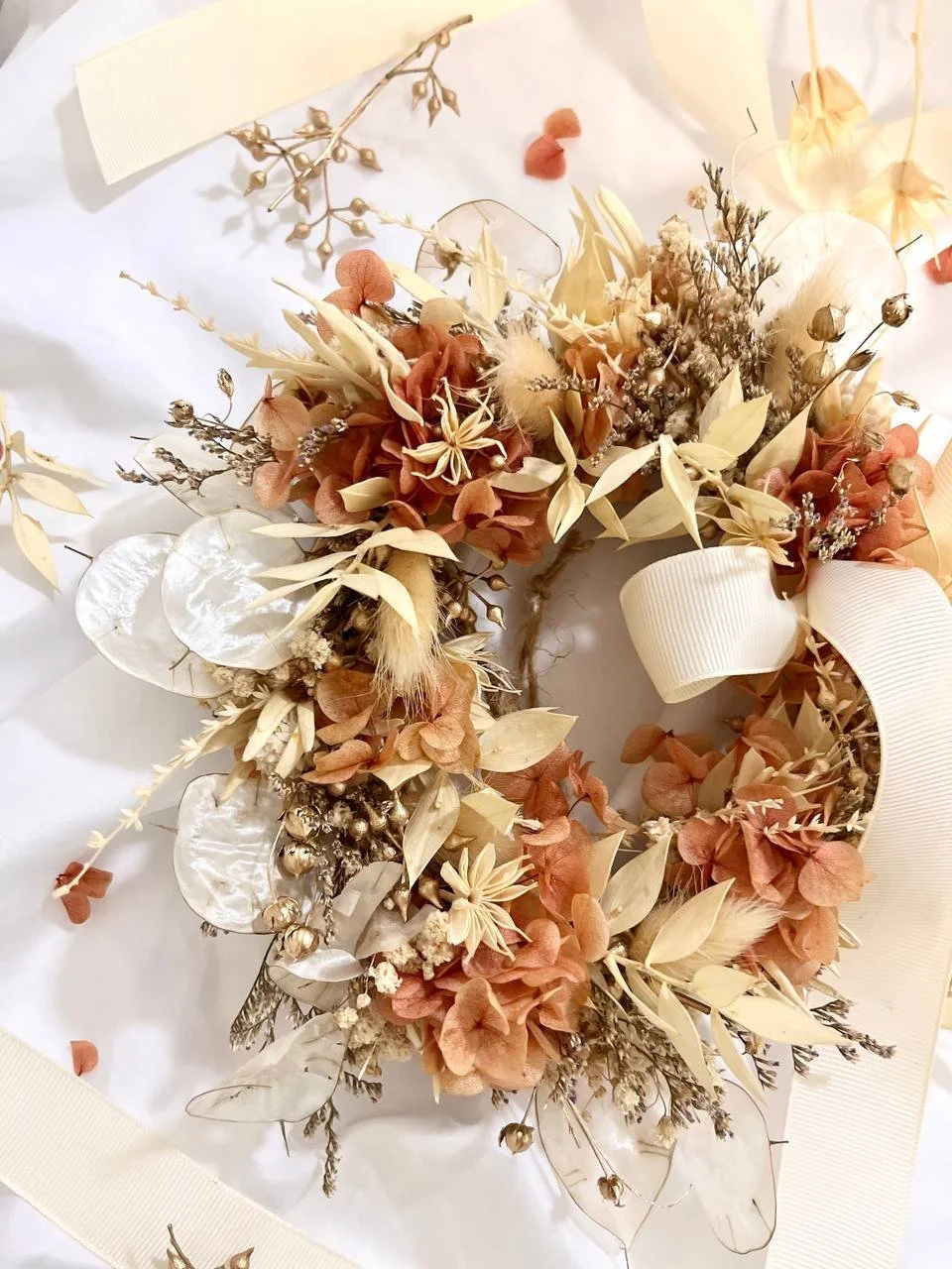 A decorative dried flower arrangement with beige, cream, and peach foliage, ribbons, and decorative elements on a white surface.