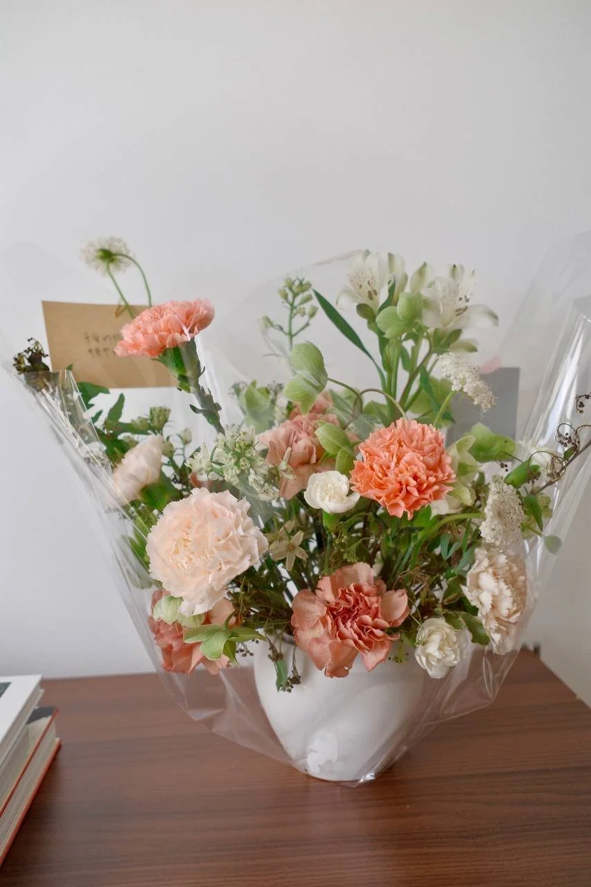 A bouquet of mixed flowers in a white vase wrapped in clear plastic on a wooden table.
