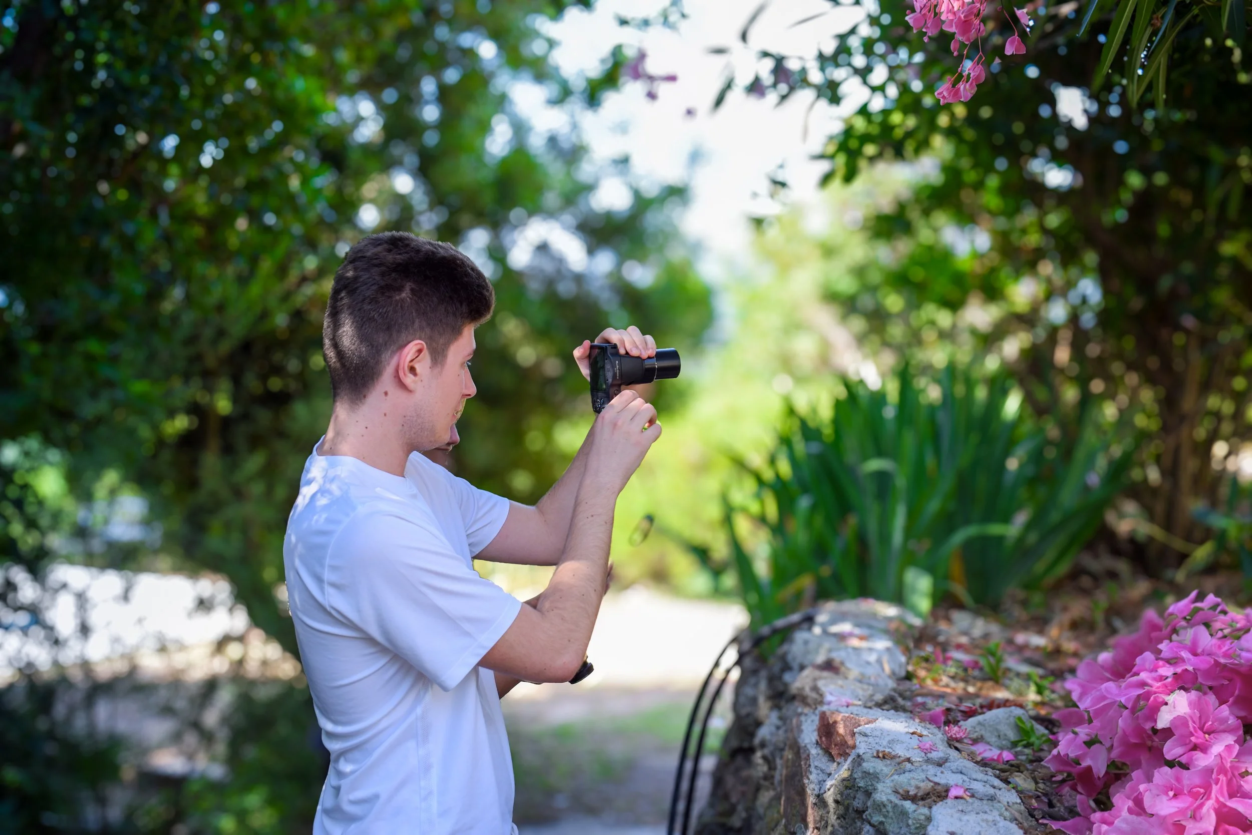 Young man in white shirt taking a photograph with a camera in a garden with green foliage and pink flowers.