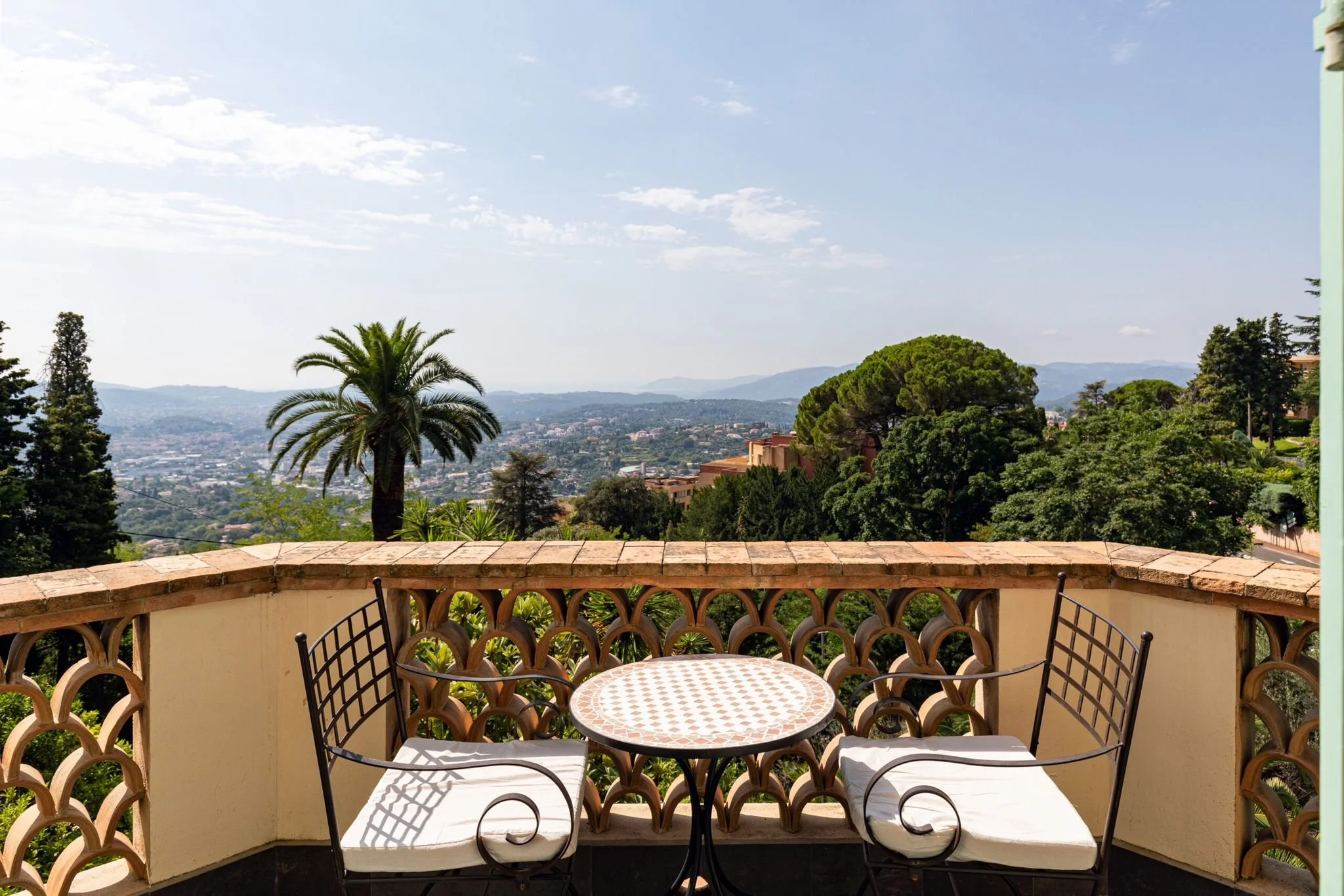 View from a balcony with two black chairs with white cushions and a small round table, overlooking a lush green landscape with trees, a palm tree, and a distant cityscape under a partly cloudy sky.