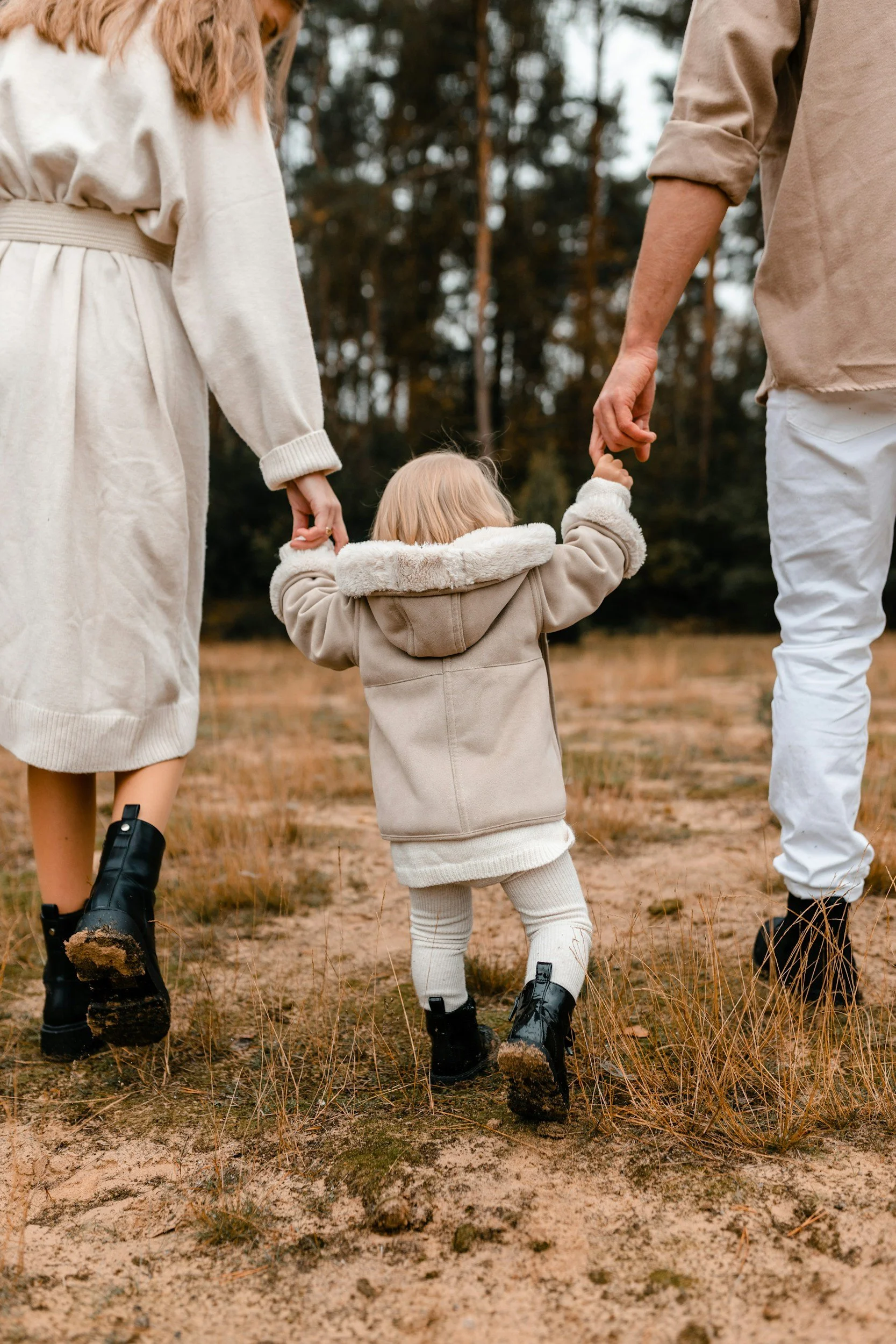 A young child dressed in warm clothing is holding hands with two adults, walking through a grassy outdoor area with trees in the background.