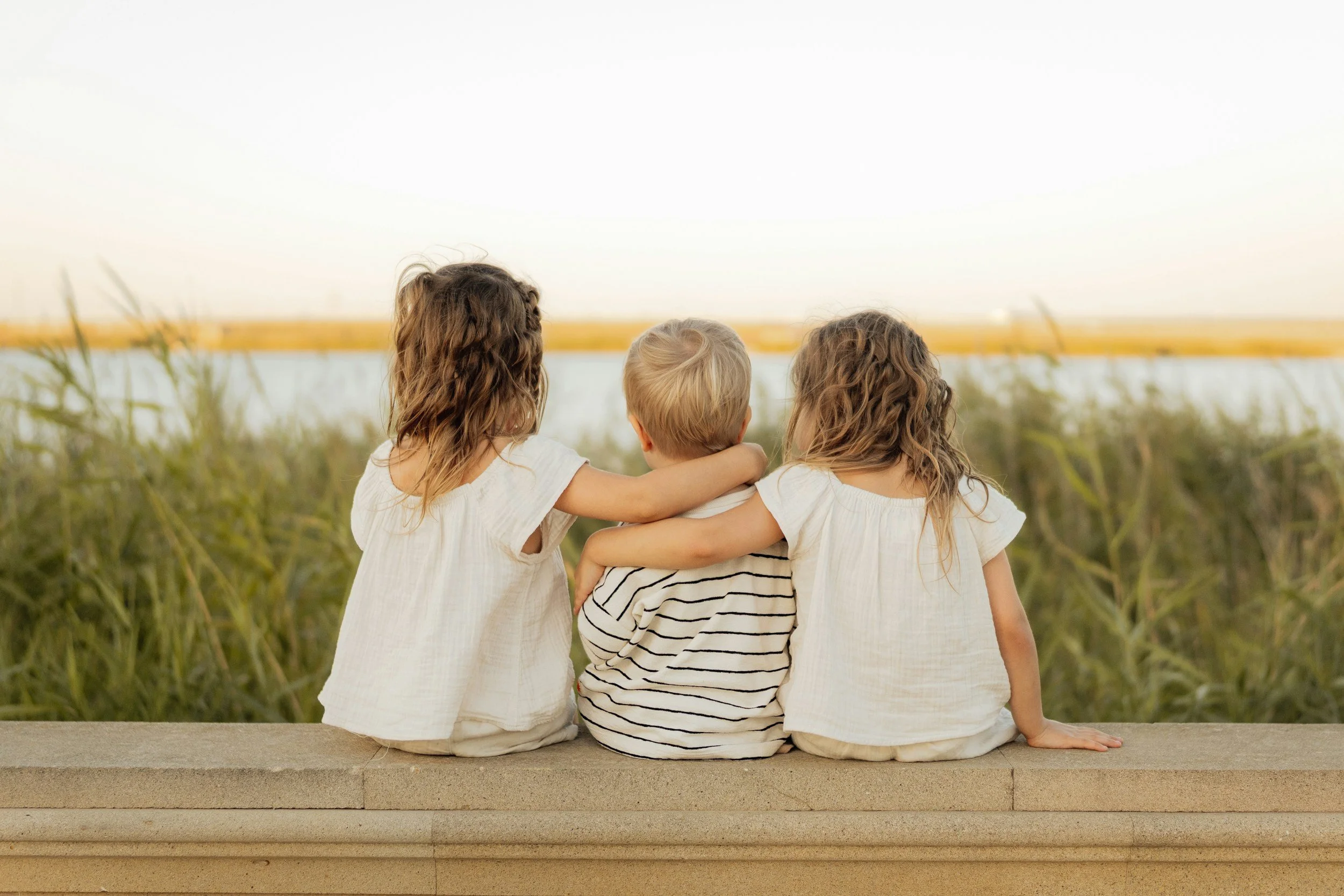 Three children sitting on a ledge, facing a body of water, with arms around each other, during sunset.